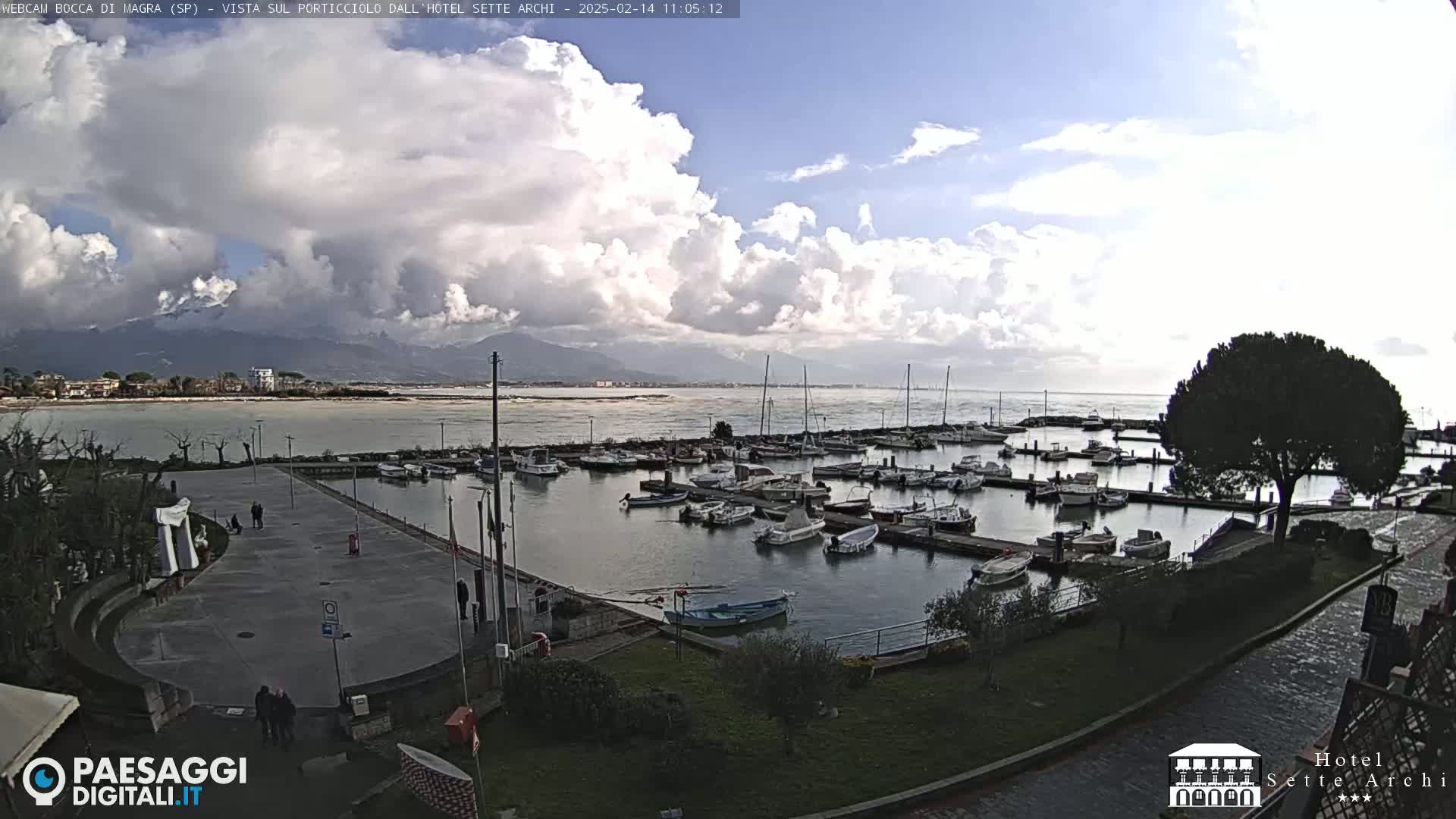 A partly cloudy day shows a marina filled with numerous boats, a paved area with a few people walking, and a view of calm water leading to mountains in the distance.