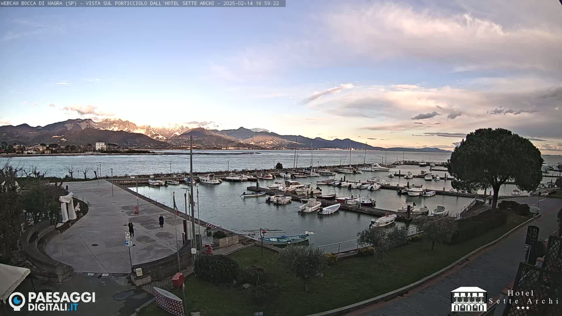 A marina filled with numerous boats is situated near a paved area with a few people, overlooking a calm body of water and mountains under a partly cloudy sky.
