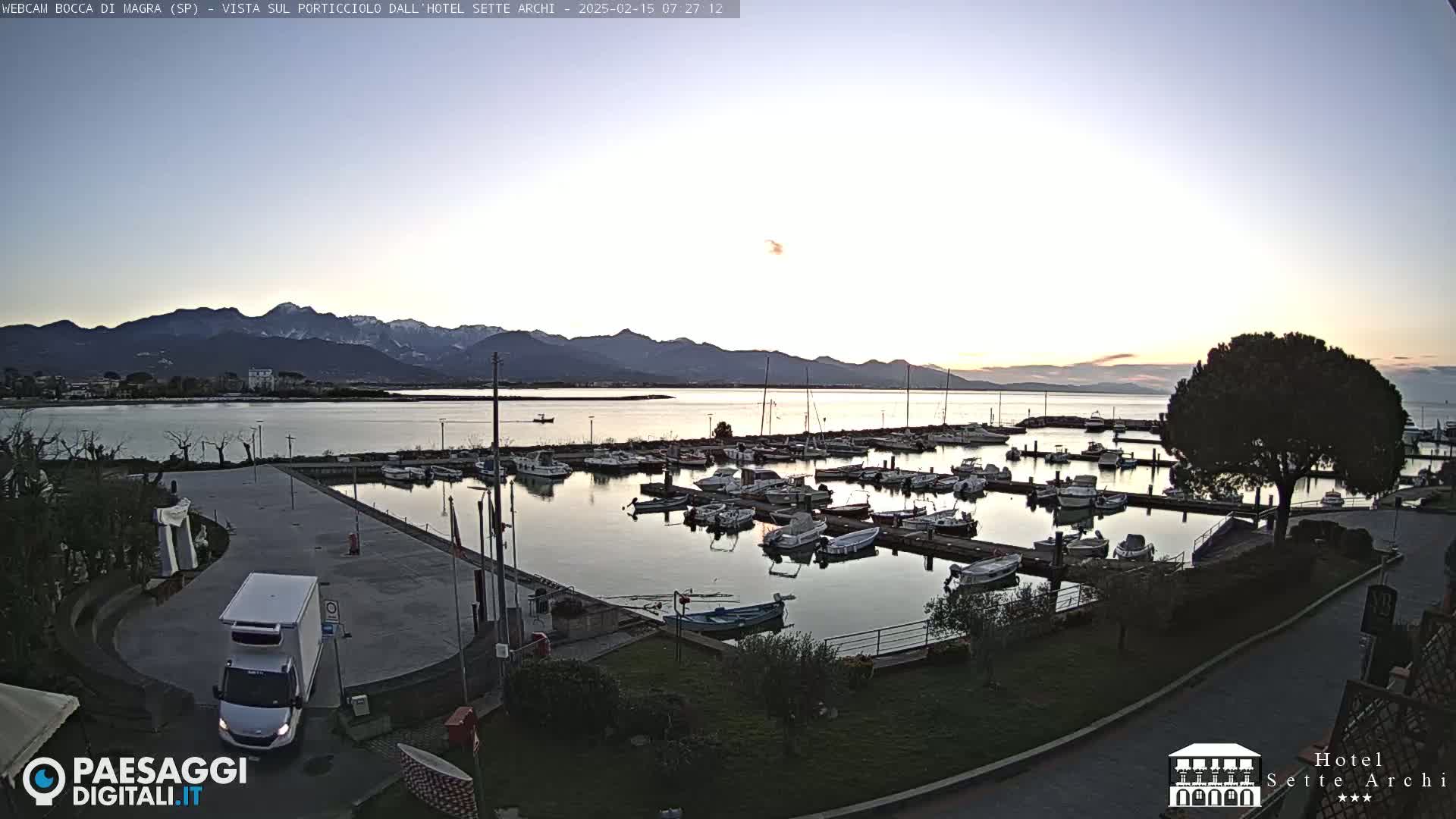 A marina filled with boats at dawn, under a pale sky, with snow-capped mountains in the distance.