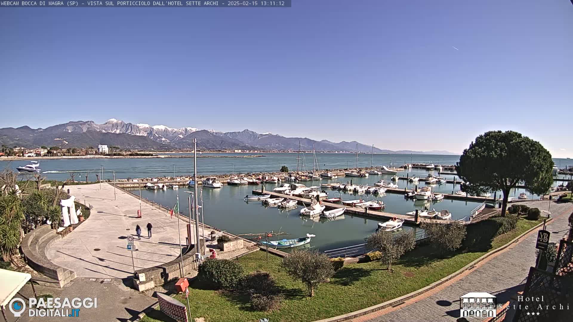 A sunny day shows a harbor filled with boats, a paved plaza with a few people walking, and snow-capped mountains in the distance.