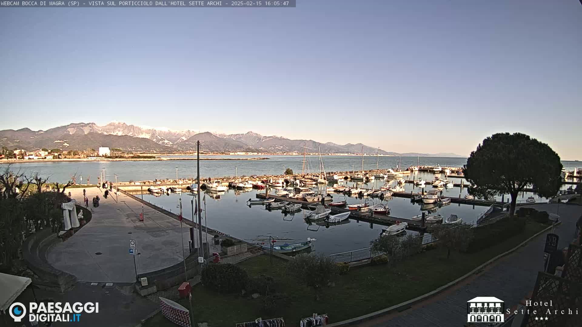 A marina filled with boats sits before a calm body of water, backed by snow-capped mountains under a clear, light-blue sky.