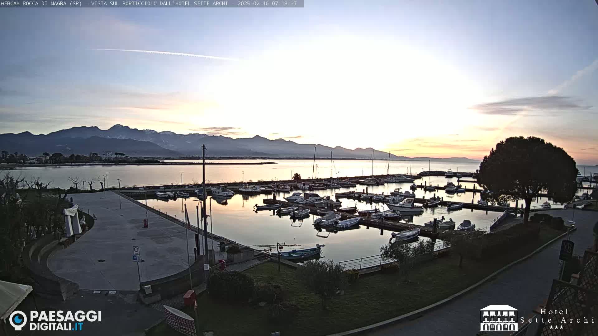 A calm harbor filled with numerous boats is seen at sunrise, with mountains in the background under a clear sky.
