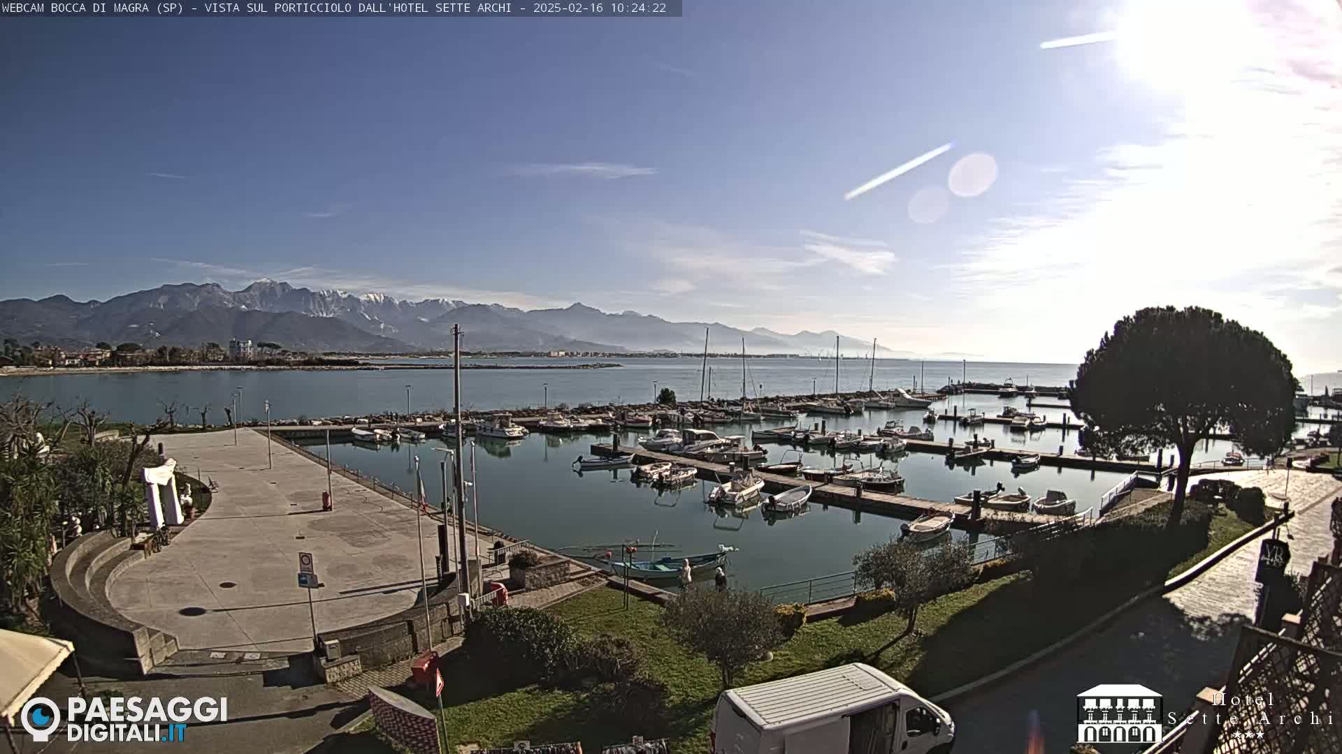 A marina filled with boats is situated near a paved area, with mountains in the background under a clear, sunny sky.