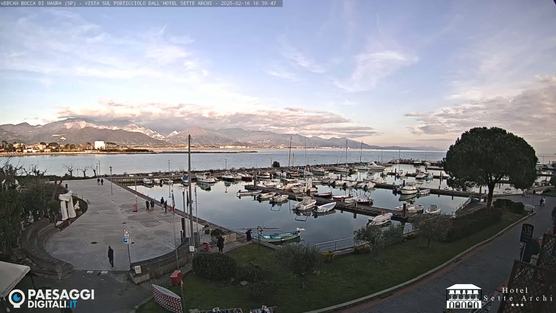 A marina filled with boats is situated near a paved walkway and green space, overlooking calm water and distant mountains under a partly cloudy sky.