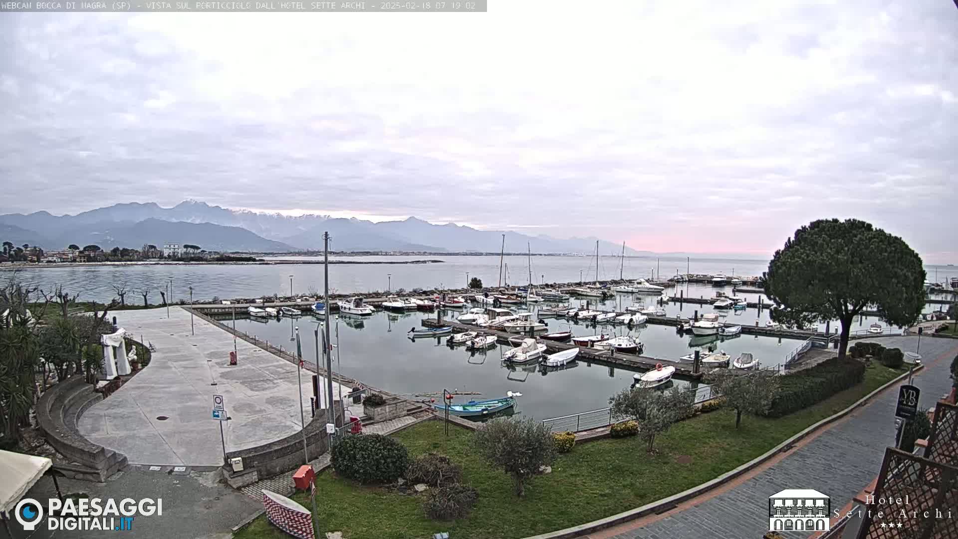 A marina filled with numerous boats is situated near a paved plaza, overlooking calm water and a mountain range under a cloudy sky.