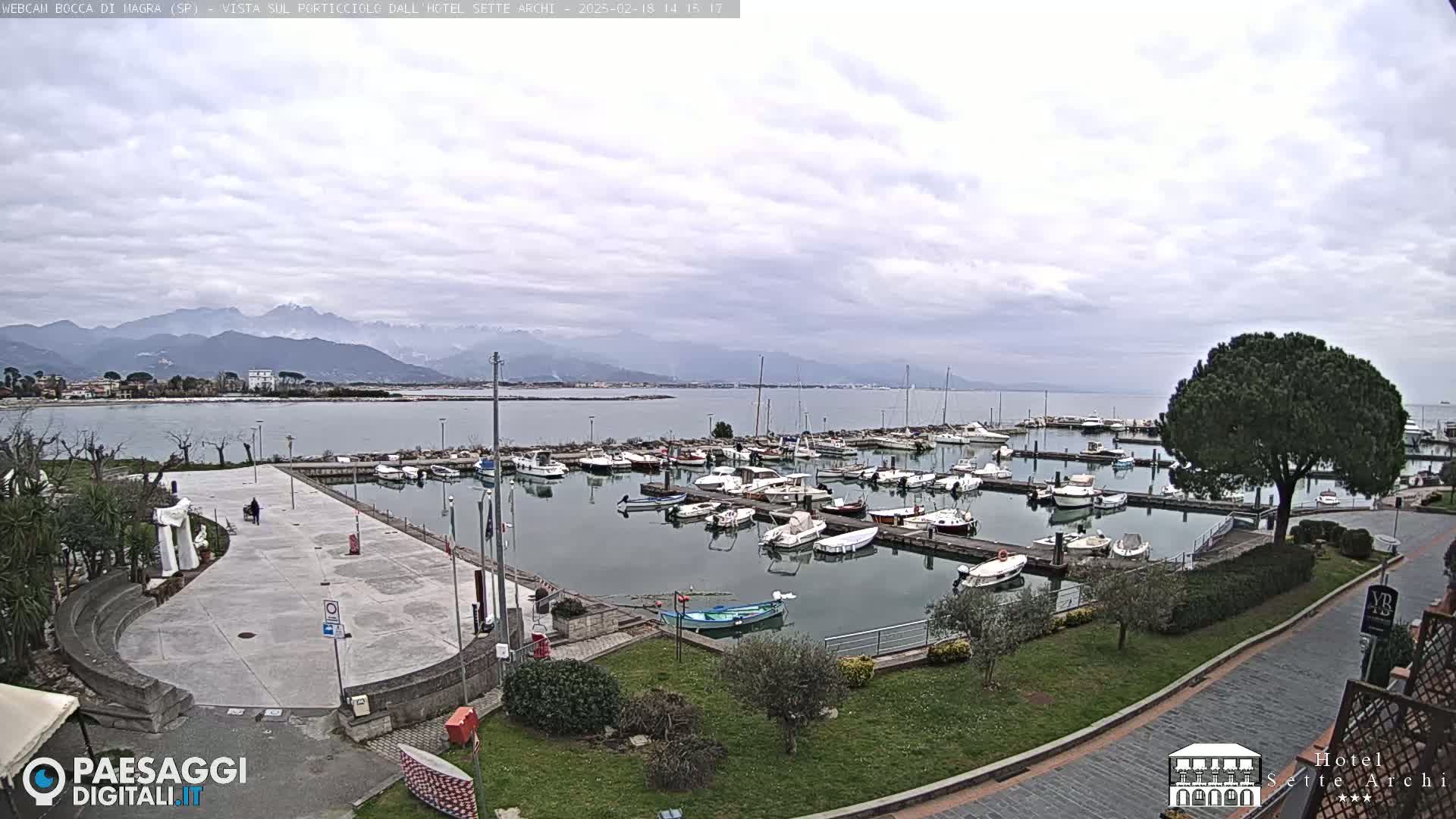 A harbor filled with boats is situated next to a paved plaza, overlooking a calm body of water and distant mountains under a cloudy sky.