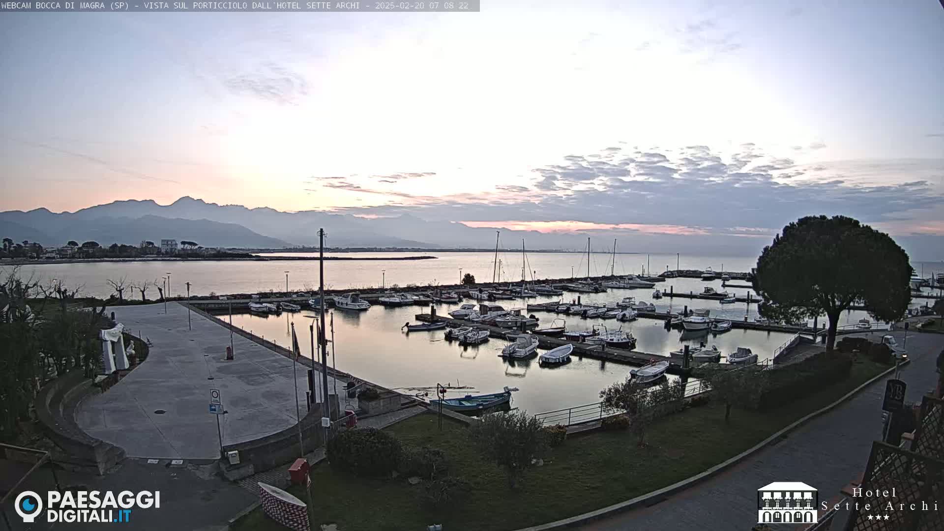 A calm marina filled with boats sits at the edge of a body of water under a pale sunrise sky with mountains in the background.