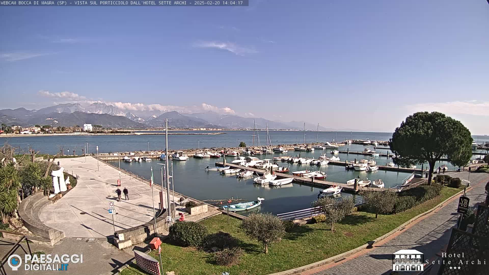 A sunny day overlooking a harbor filled with boats, a paved plaza, and snow-capped mountains in the distance.