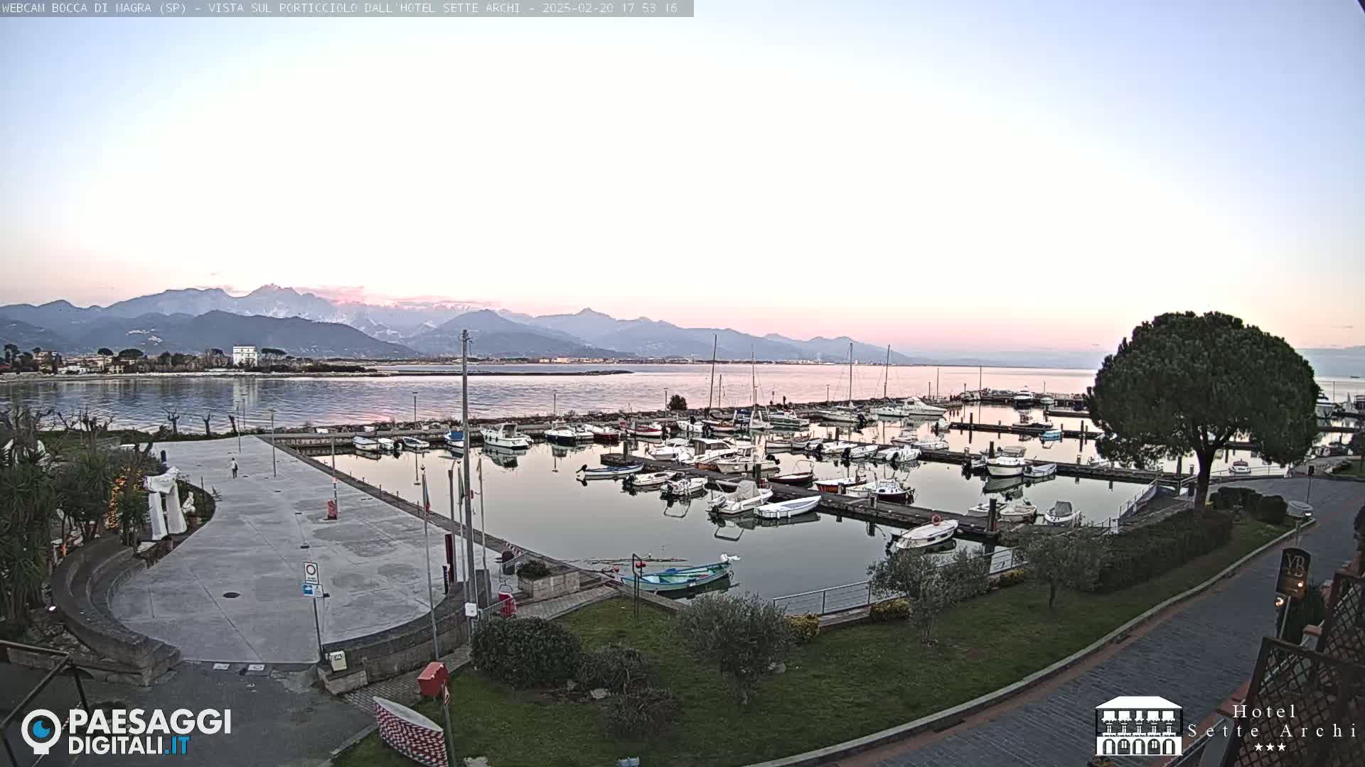 A harbor filled with numerous boats is situated next to a paved plaza, all overlooking a calm body of water with snow-capped mountains in the distance under a pale, twilight sky.