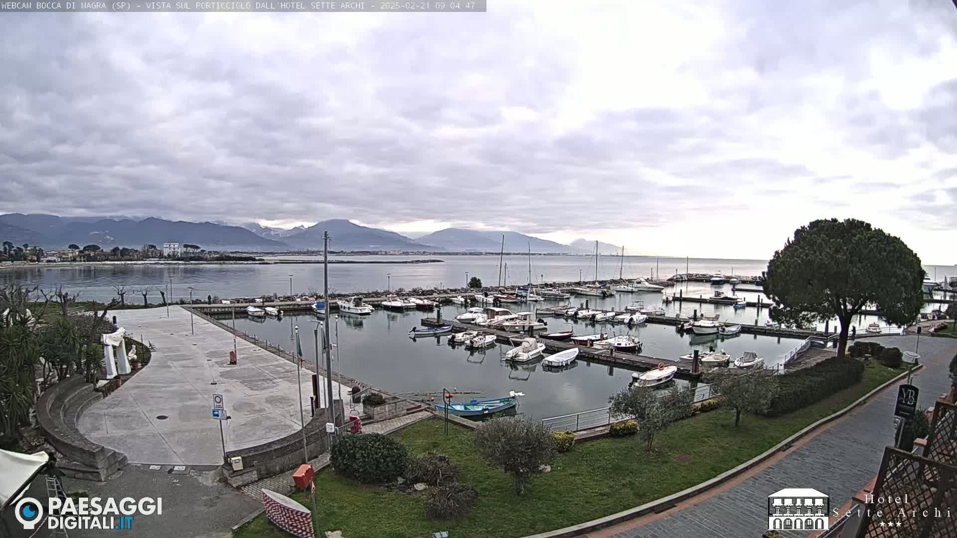 A marina filled with boats sits beside a paved plaza, overlooking calm water and distant mountains under a cloudy sky.