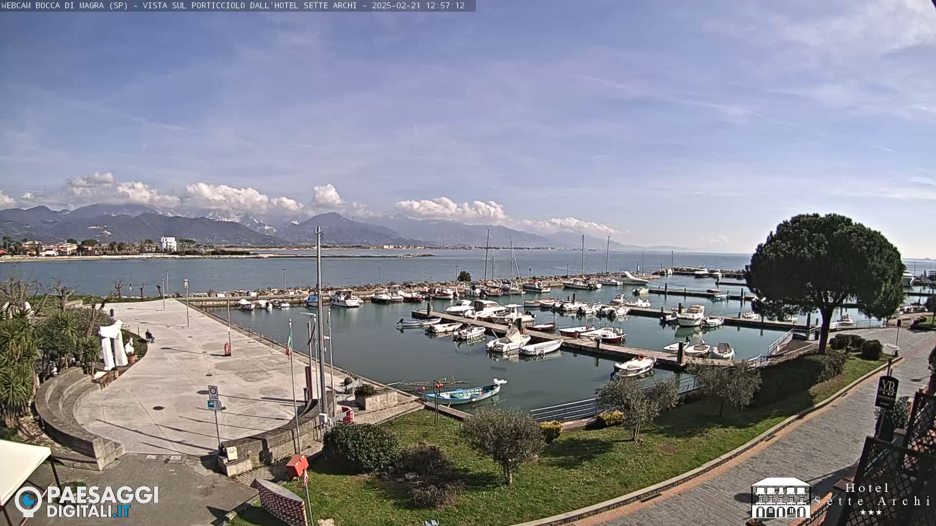A marina filled with boats sits beside a paved plaza and green landscaping, all under a partly cloudy sky with mountains in the distance.