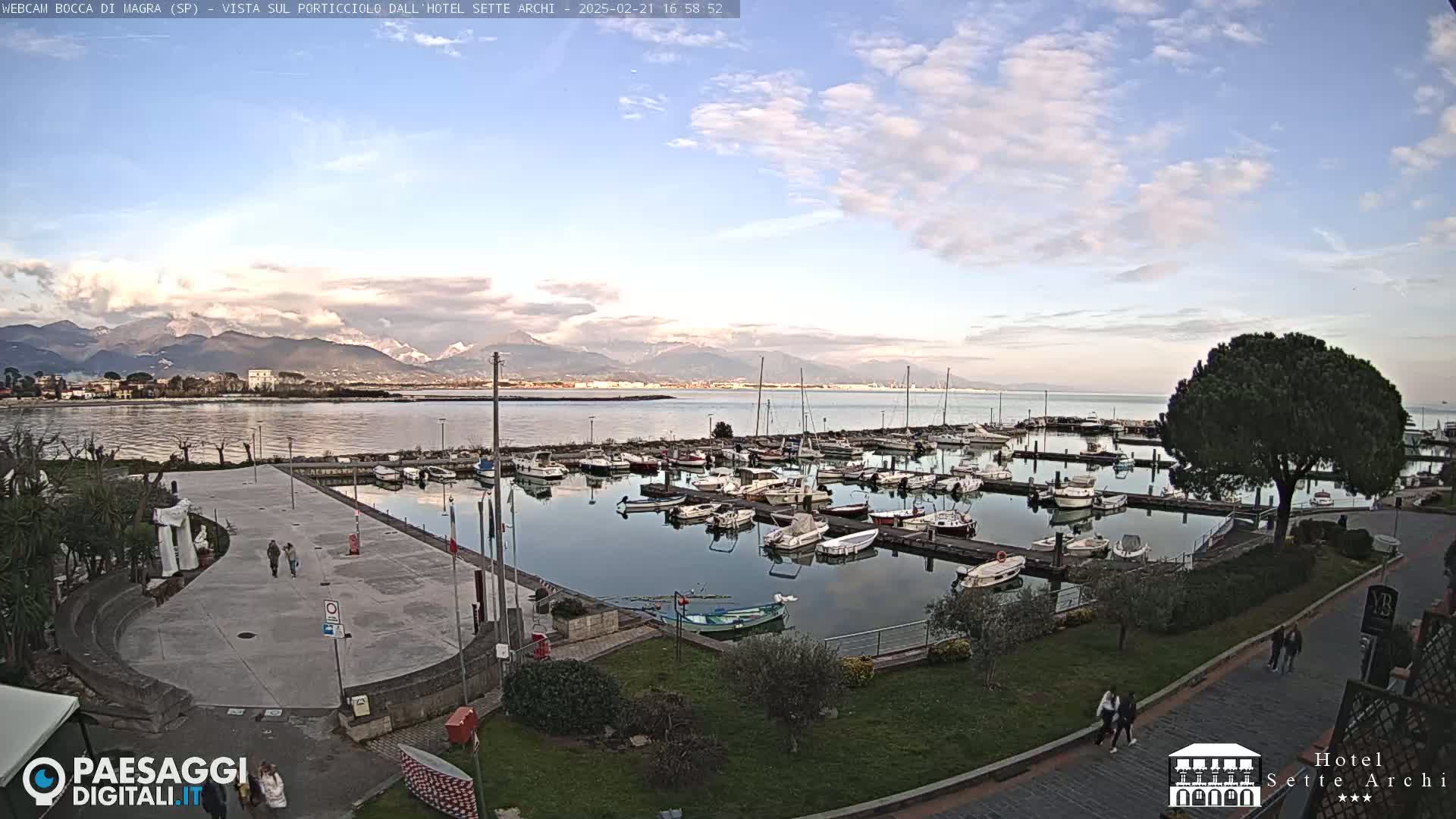 A marina filled with boats is situated next to a paved plaza and green space, overlooking a calm body of water with mountains in the distance under a partly cloudy sky.