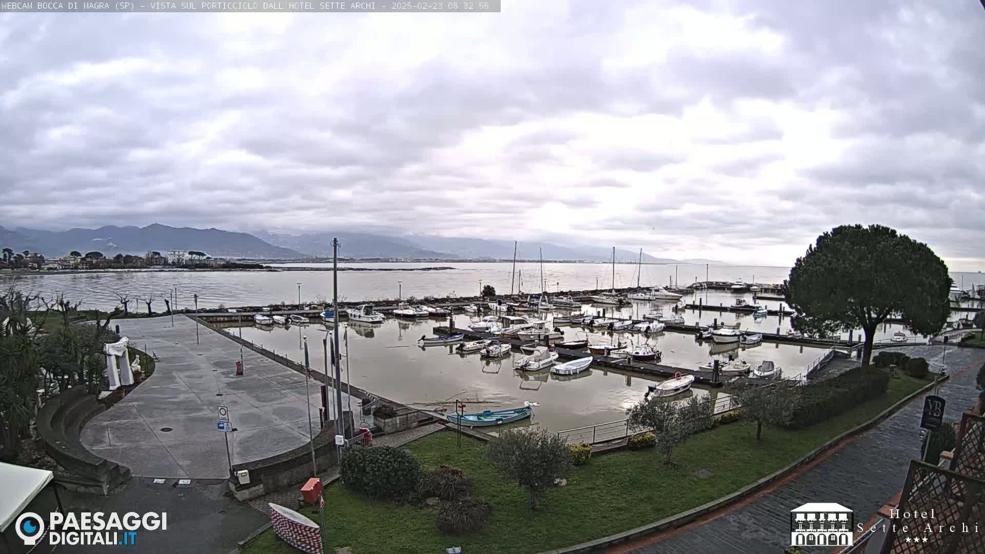 A marina filled with numerous boats is shown under a cloudy sky, with mountains visible in the distance.
