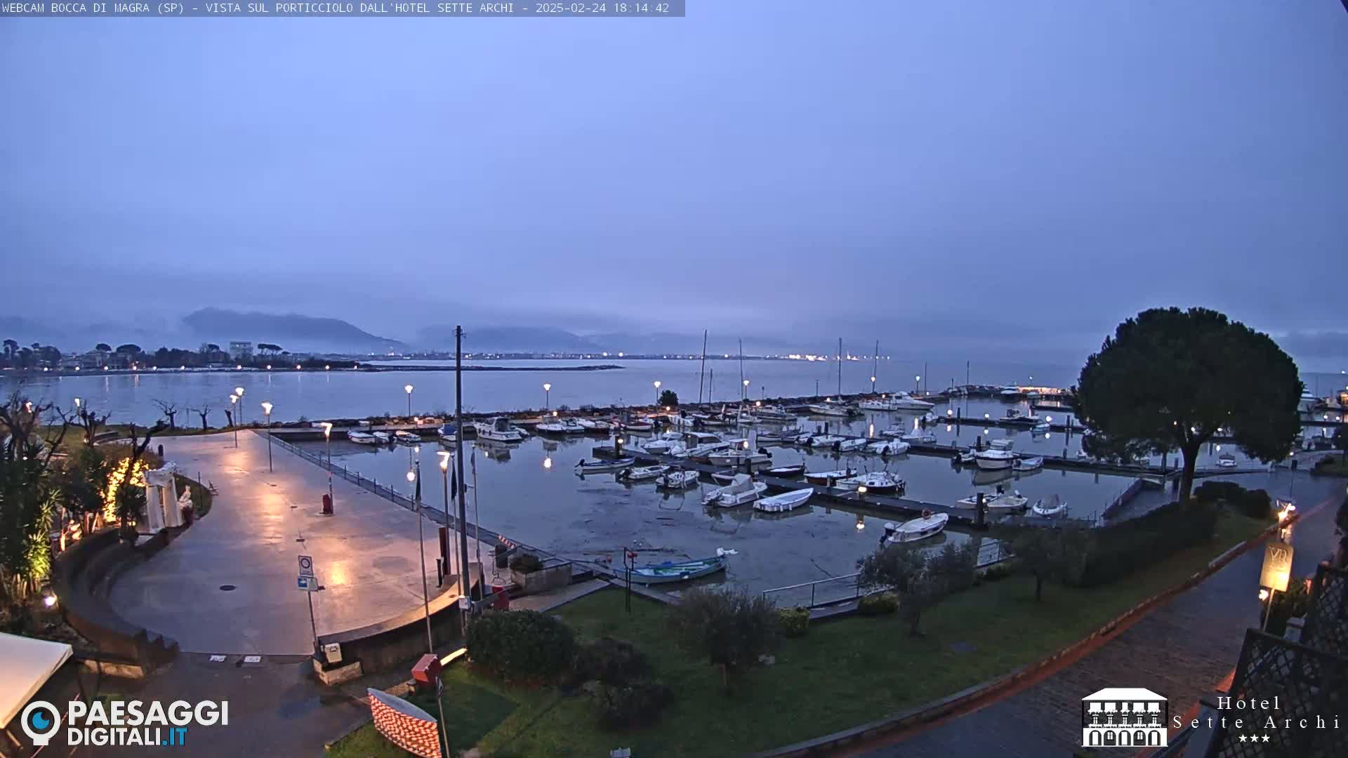A marina filled with boats at dusk, under a clear, twilight sky, overlooks a calm body of water with distant mountains.