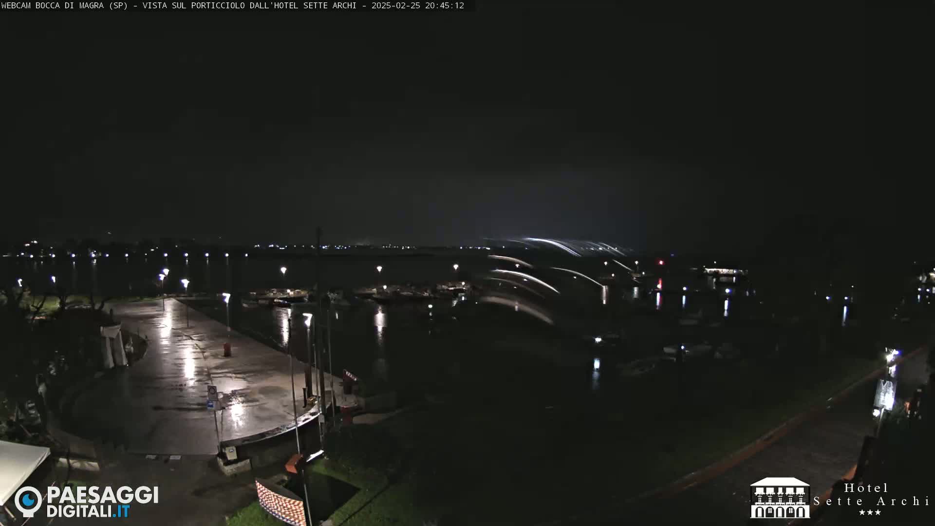 A nighttime view of a wet, paved waterfront area with boats in a harbor under a dark sky with streaks of light.