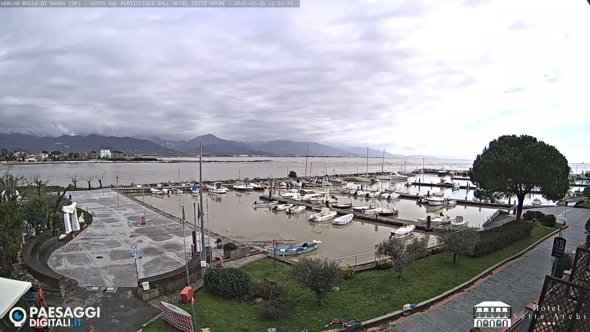 A harbor filled with boats sits under a cloudy sky, with mountains visible in the distance.