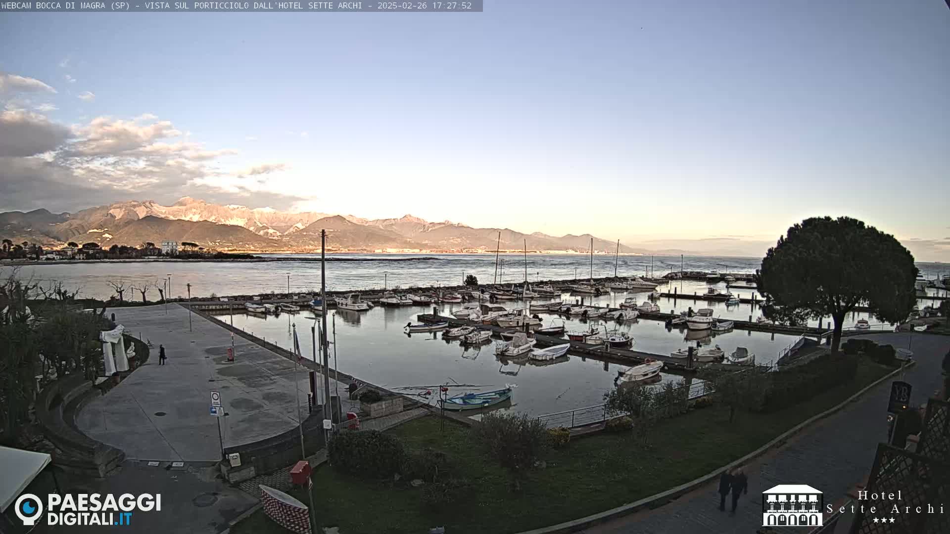 A marina filled with boats sits beside a paved area overlooking calm water and snow-capped mountains under a mostly clear sky.