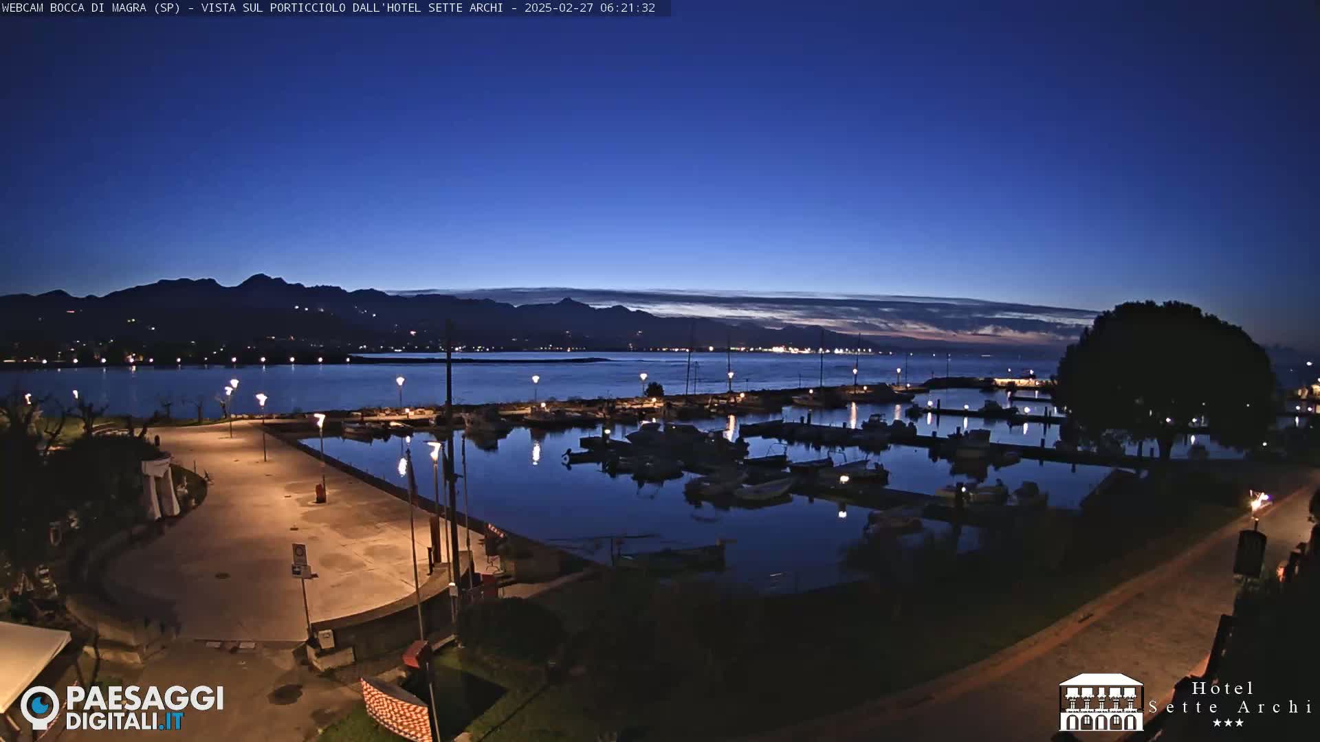 A marina filled with boats is illuminated at twilight under a clear, dark blue sky, with mountains visible in the distance.