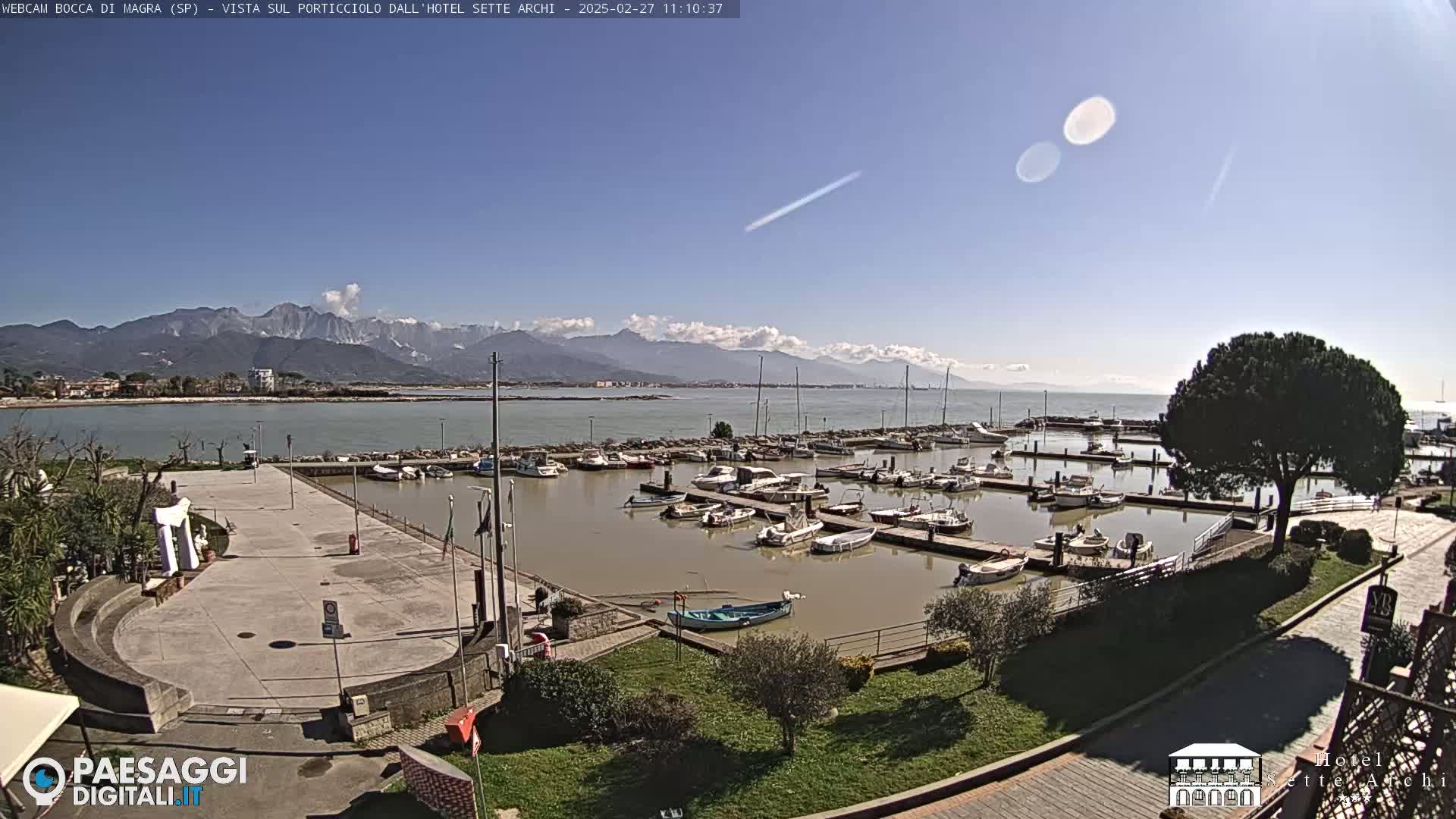A marina filled with boats is situated next to a paved plaza, all under a mostly clear sky with mountains visible in the distance.