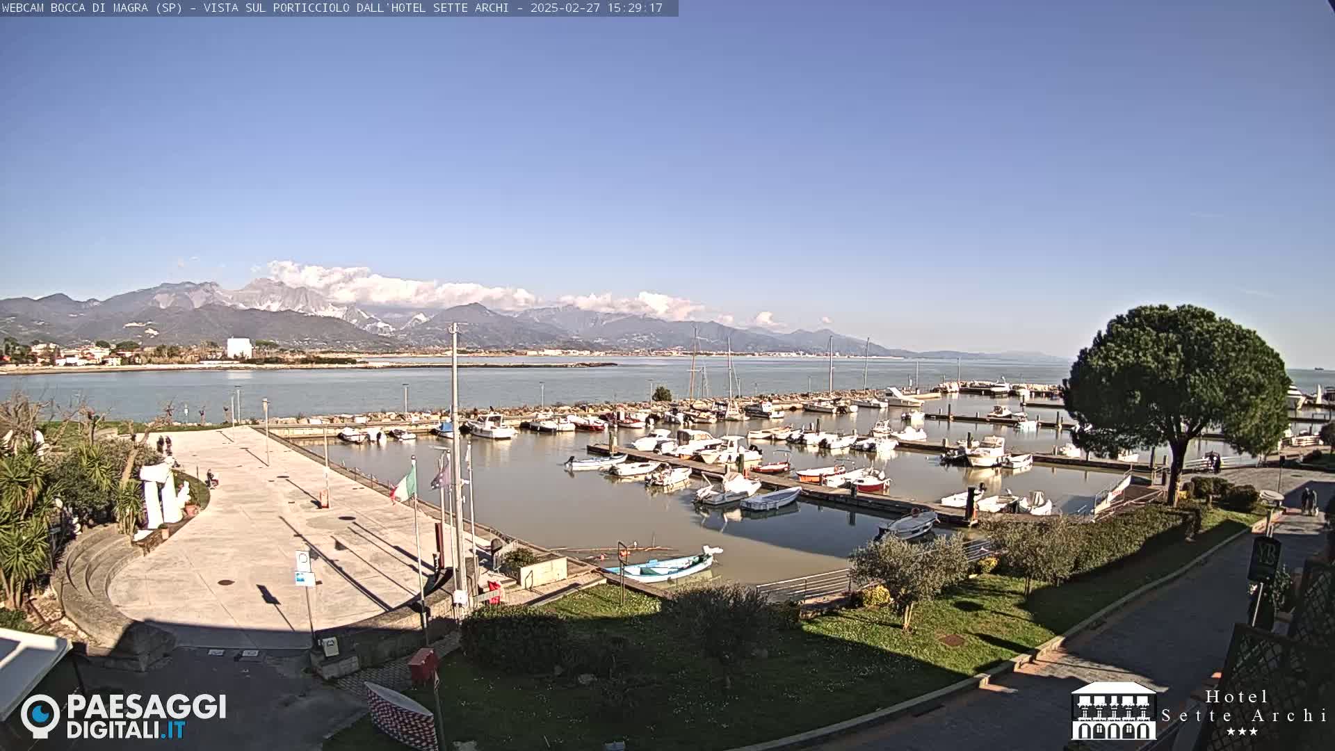 A marina filled with numerous boats is situated near a paved plaza, overlooking a calm body of water and a mountain range under a clear, sunny sky.