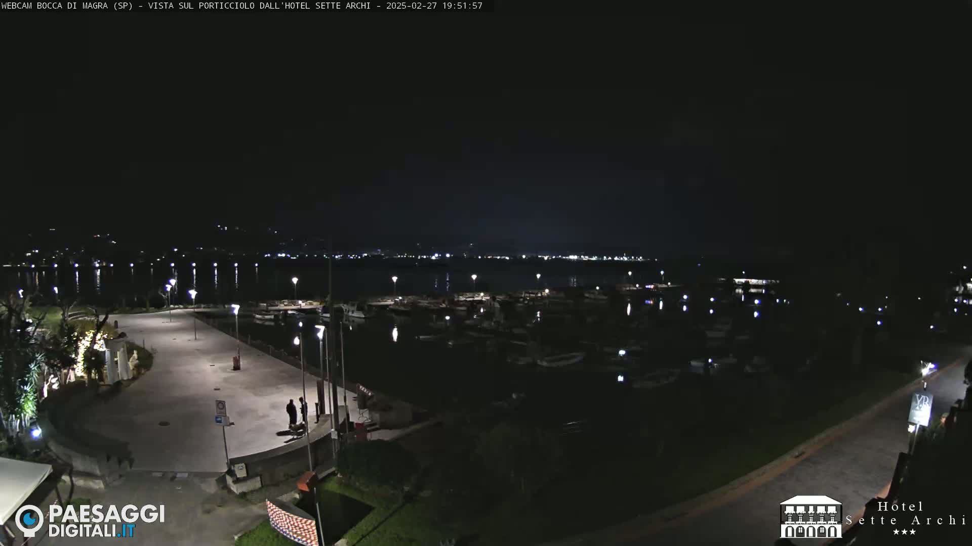 A nighttime view of a harbor filled with boats, a paved waterfront area with a few people, and a dark body of water extending to a distant, dimly lit shoreline.