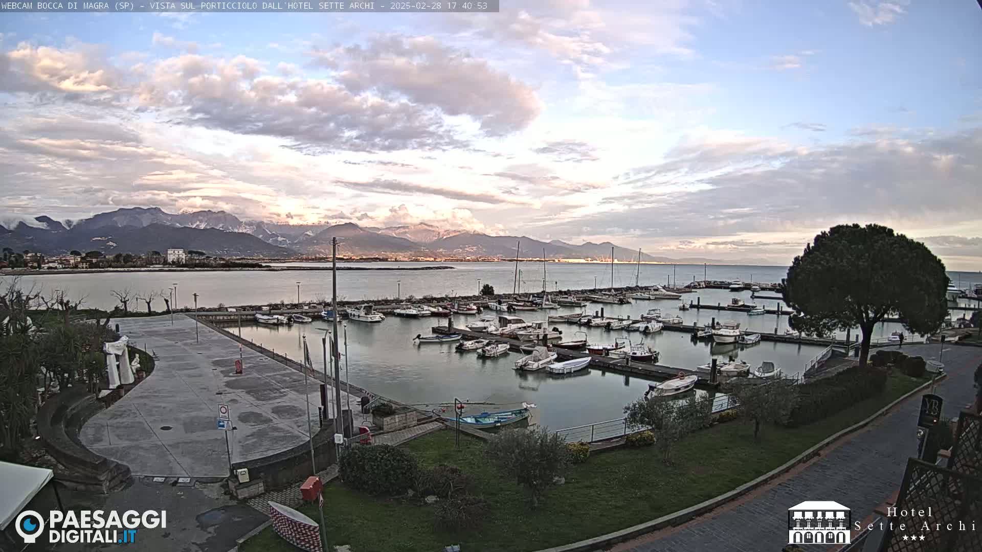 A marina filled with boats is situated next to a paved area, overlooking calm water and snow-capped mountains under a partly cloudy sky.