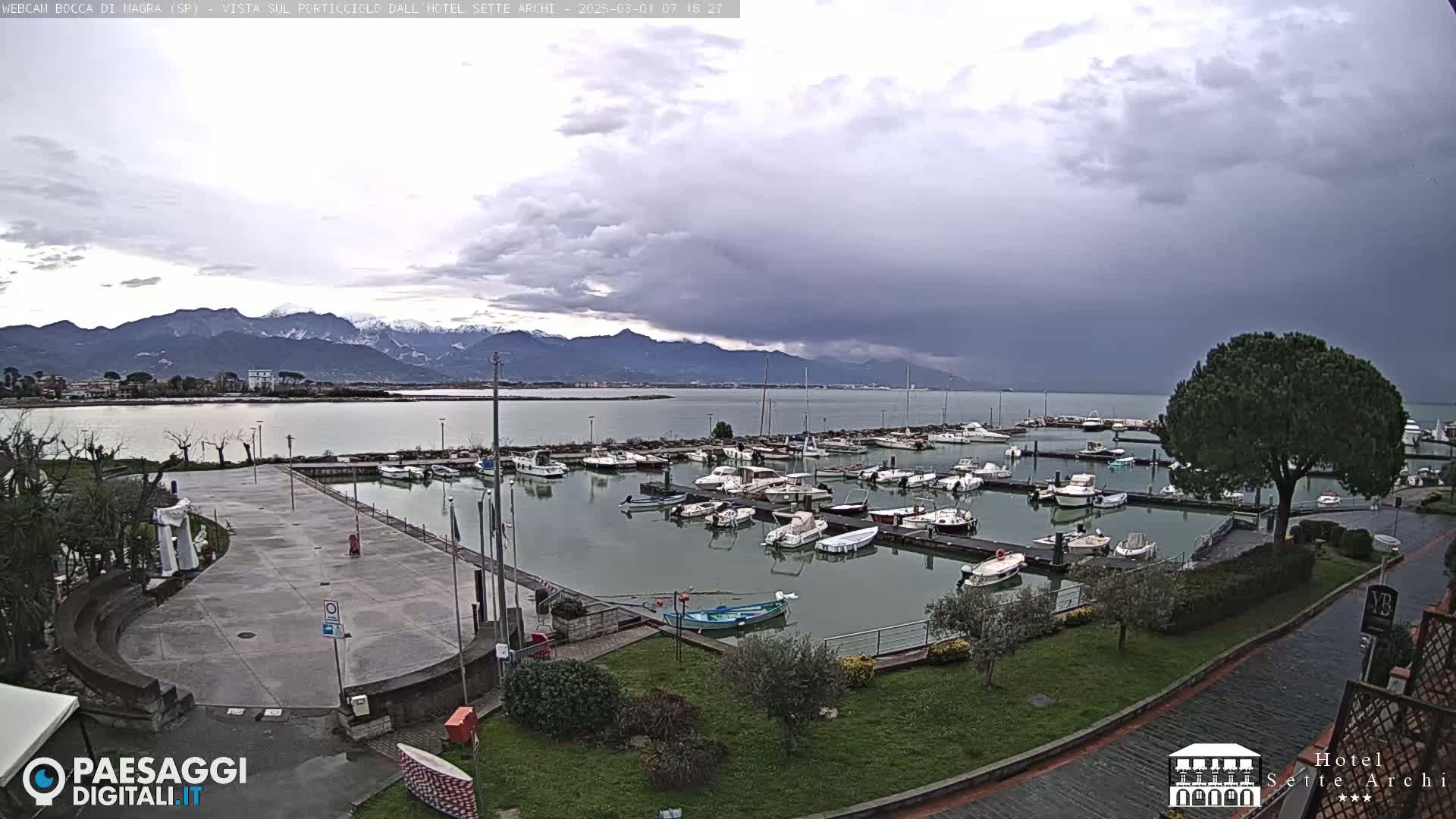 A harbor filled with numerous boats is shown under an overcast sky, with snow-capped mountains visible in the distance.