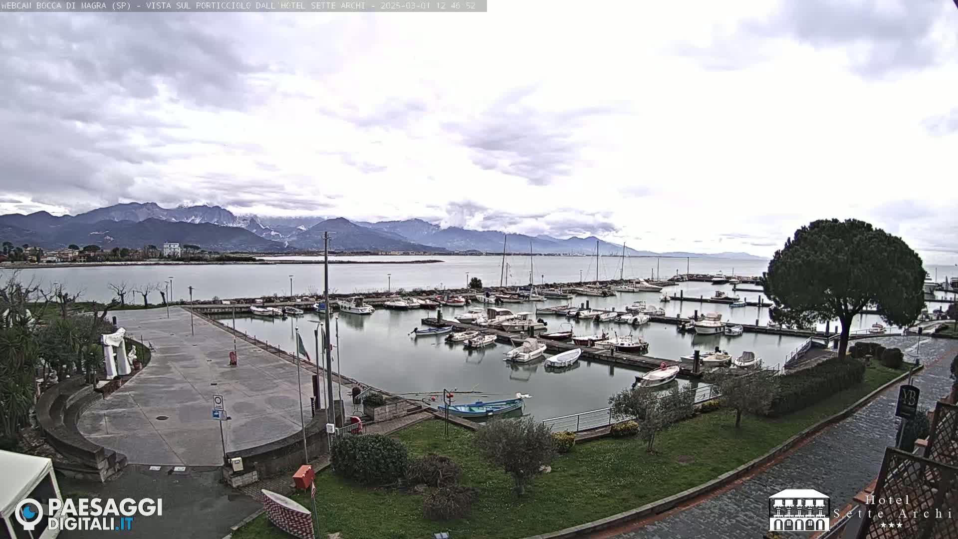 A marina filled with boats is situated near a paved area, with a mountain range in the background under a cloudy sky.
