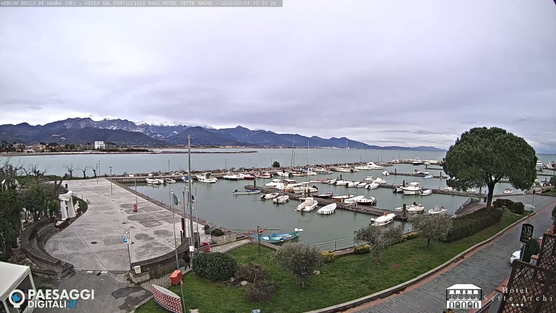 A marina filled with boats is situated next to a paved plaza, overlooking a calm body of water with snow-capped mountains in the distance under a cloudy sky.