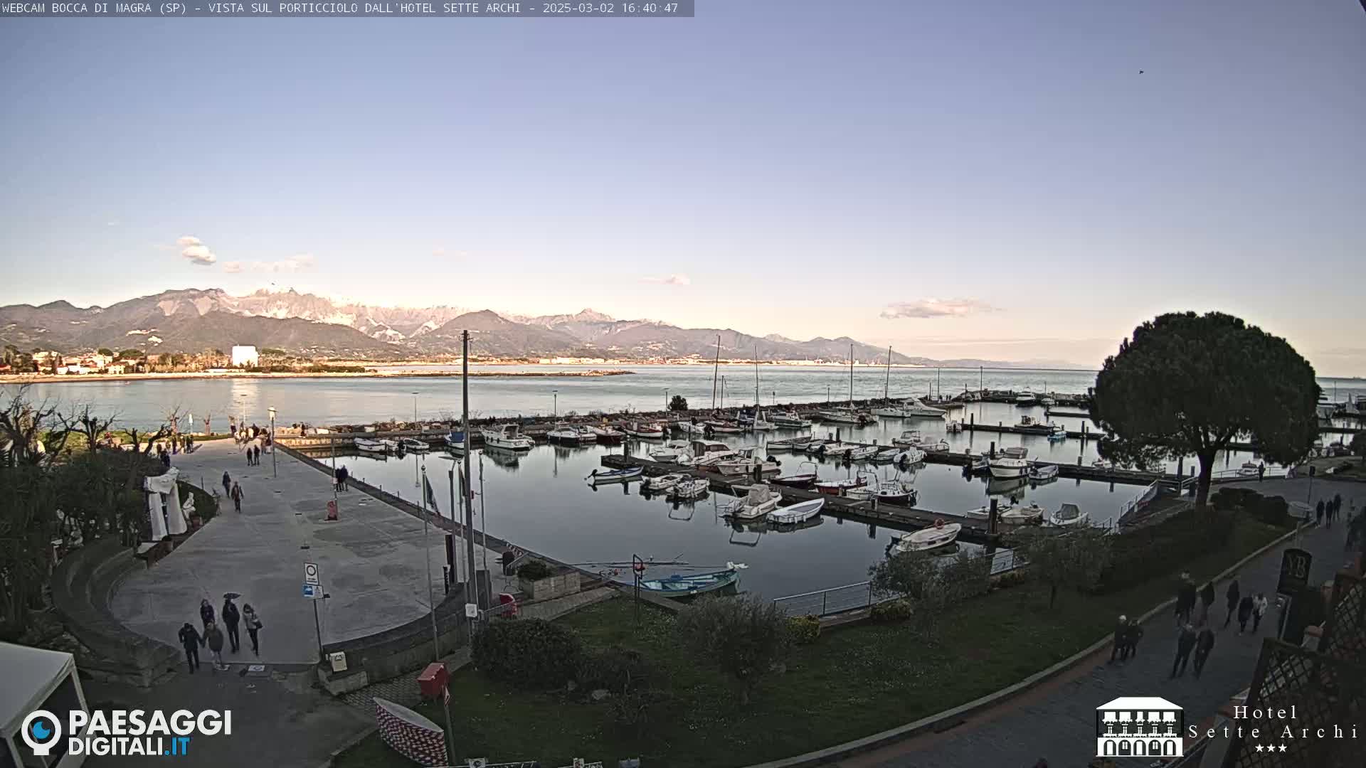 A marina filled with boats sits beside a paved walkway with people strolling, all overlooking calm water and distant mountains under a clear, twilight sky.