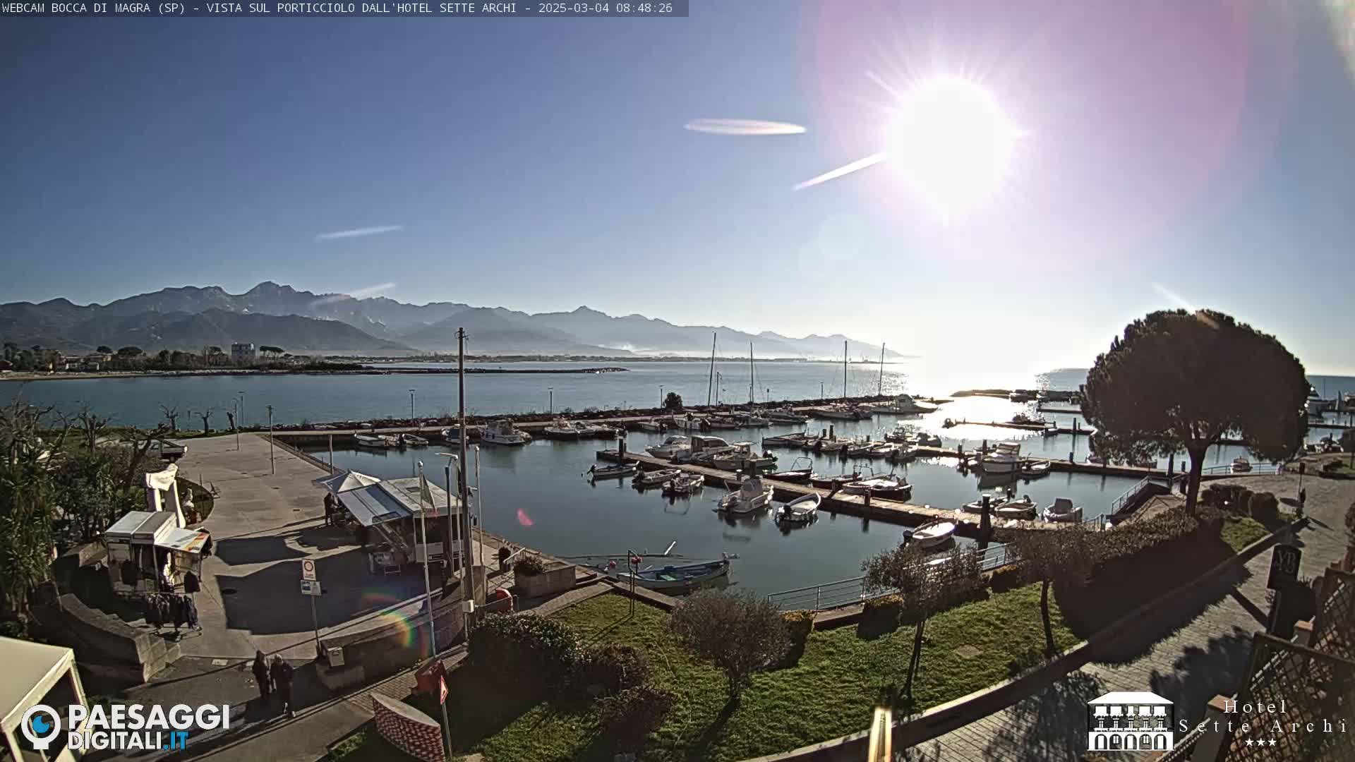 A sunny day overlooks a marina filled with boats, a paved area with small structures, and distant mountains.
