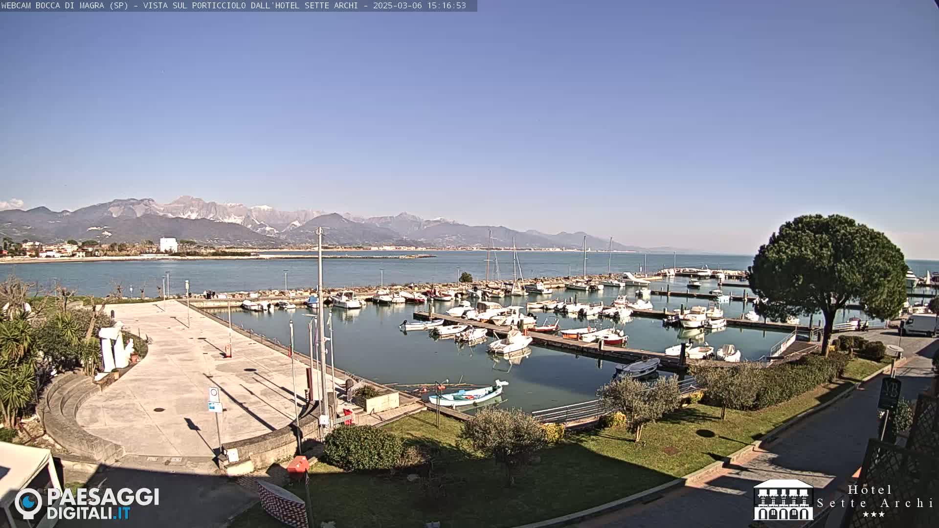 A marina filled with boats is situated beside a paved area, overlooking calm water and mountains in the distance under a clear blue sky.