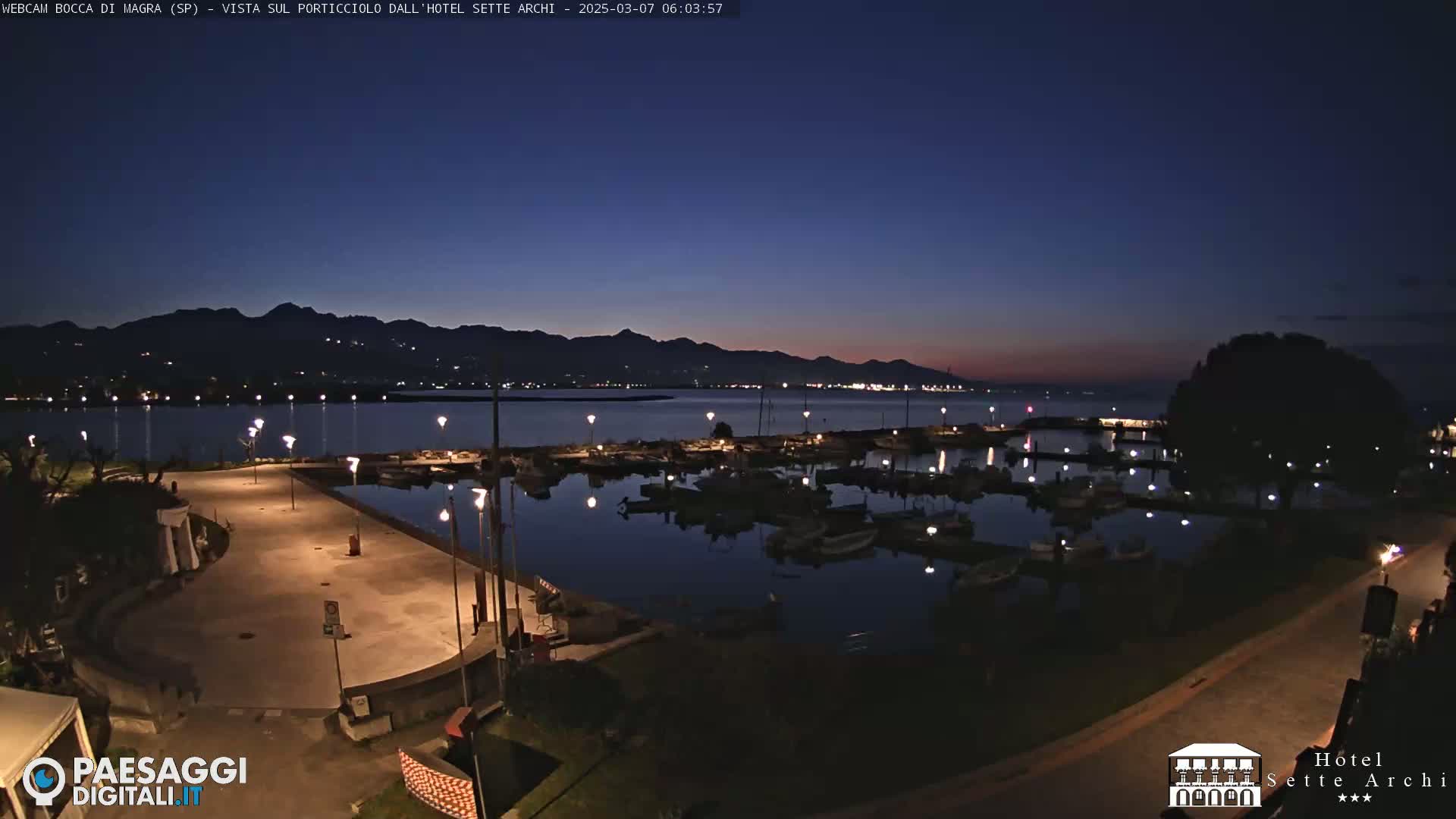 A nighttime view of a marina filled with boats, illuminated by lights, sits before a dark, mountainous shoreline under a twilight sky.
