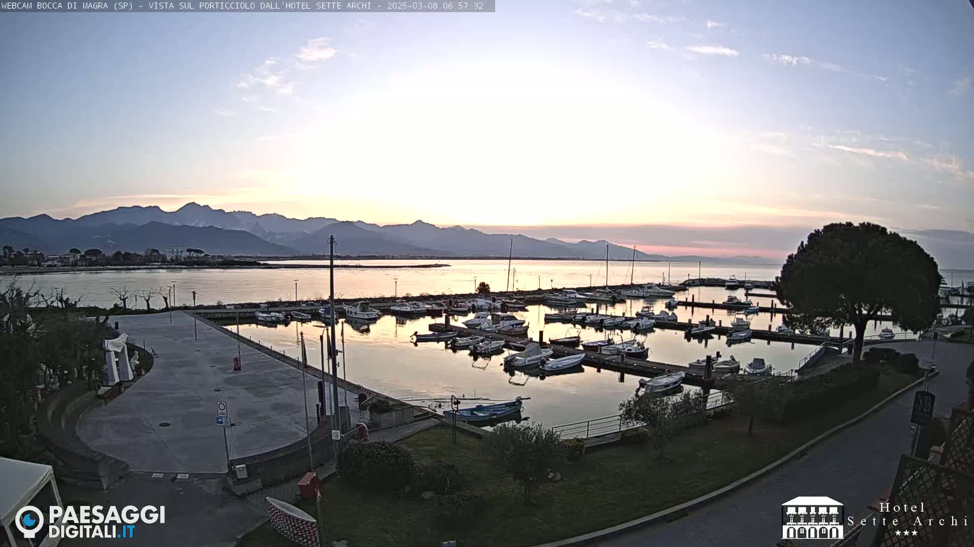 A calm harbor filled with boats at sunrise, backed by mountains under a mostly clear sky.