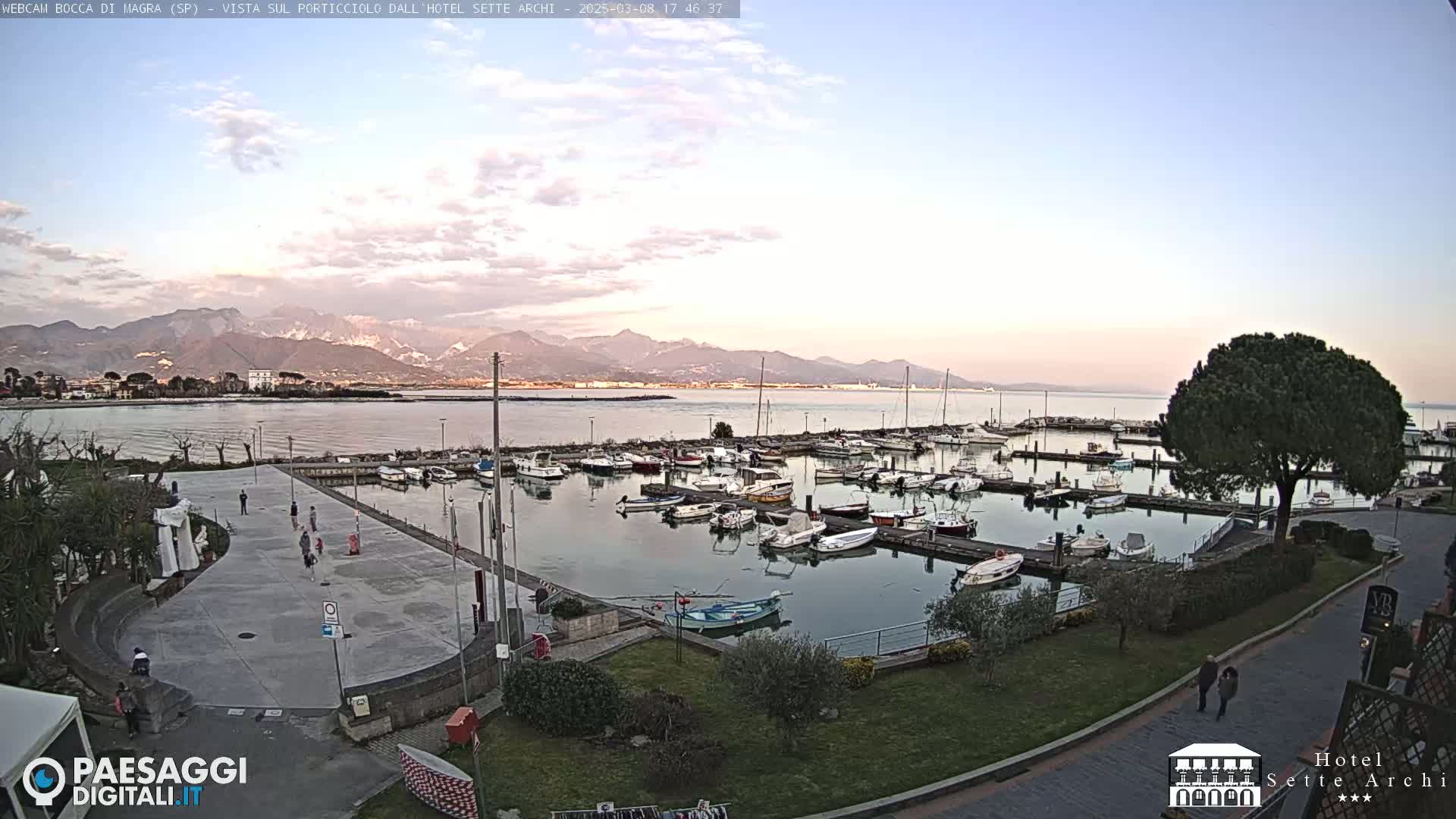 A calm body of water with a marina full of boats is seen under a mostly clear sky, with mountains visible in the distance.