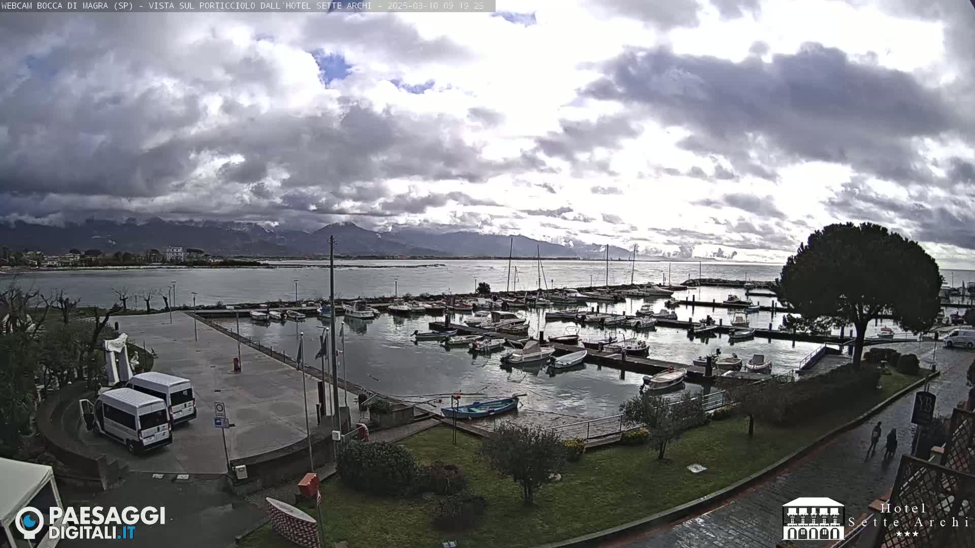 A harbor filled with numerous boats is seen under a cloudy sky, with mountains in the background.