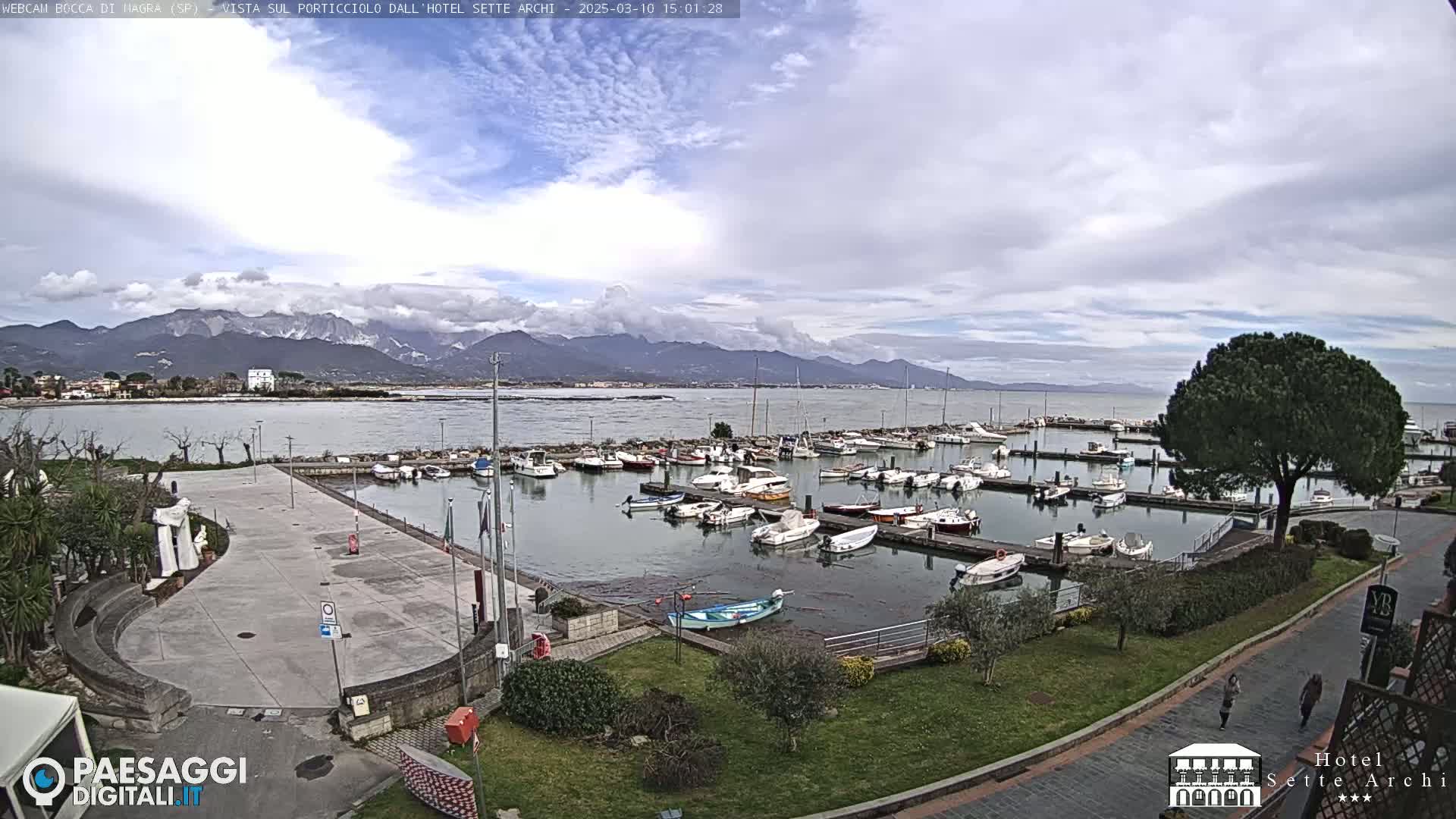 A harbor filled with numerous boats is situated near a paved plaza and lush green space, all set against a backdrop of mountains under a partly cloudy sky.