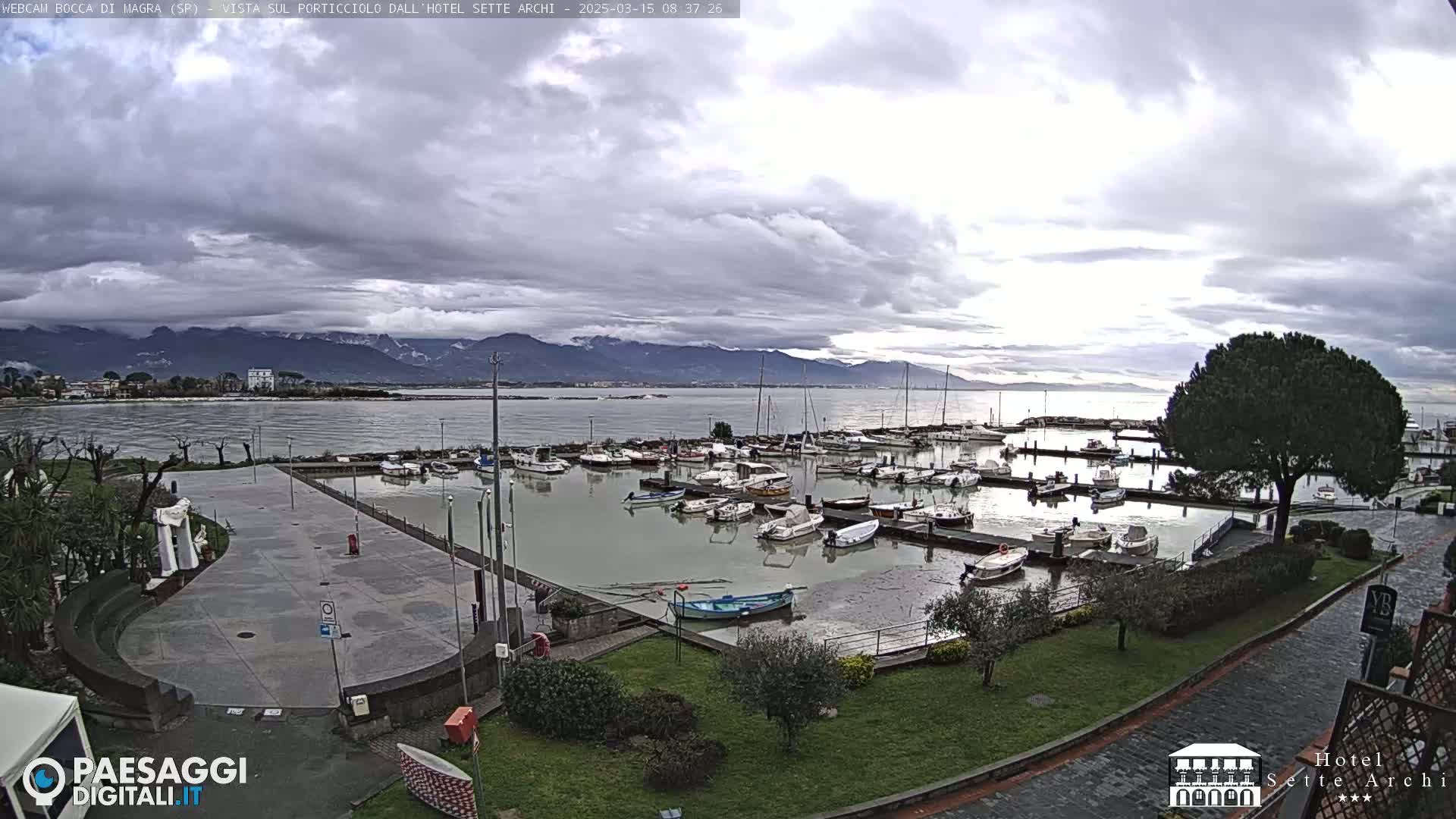 A marina filled with boats sits next to a paved plaza, overlooking a calm body of water with mountains in the distance under a cloudy sky.