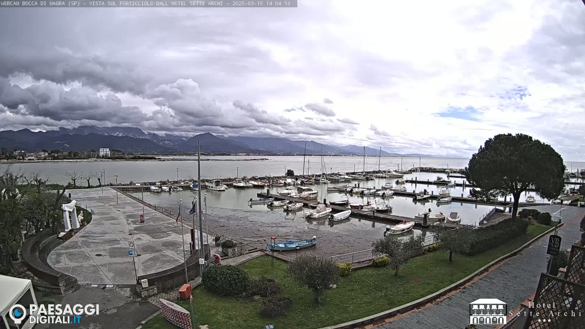 A harbor filled with docked boats is shown under a cloudy sky, with mountains in the distance.