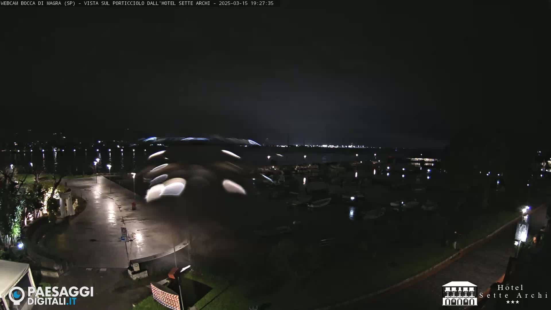 A nighttime view of a wet, paved waterfront area with boats in a marina and lights across the water.