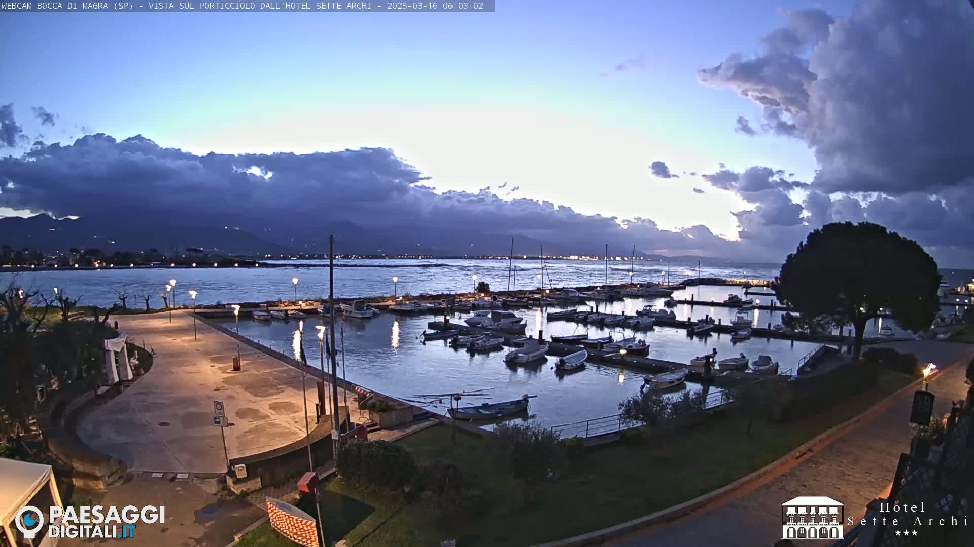 A marina filled with boats is situated on a calm body of water under a partly cloudy twilight sky, with mountains visible in the distance.