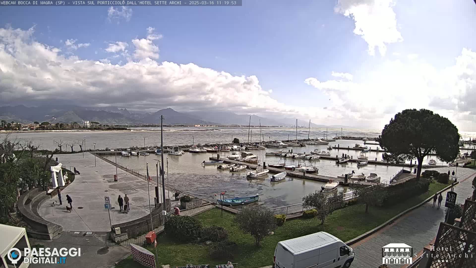 A partly cloudy day reveals a marina filled with boats, a paved plaza with pedestrians, and distant mountains across a body of water.