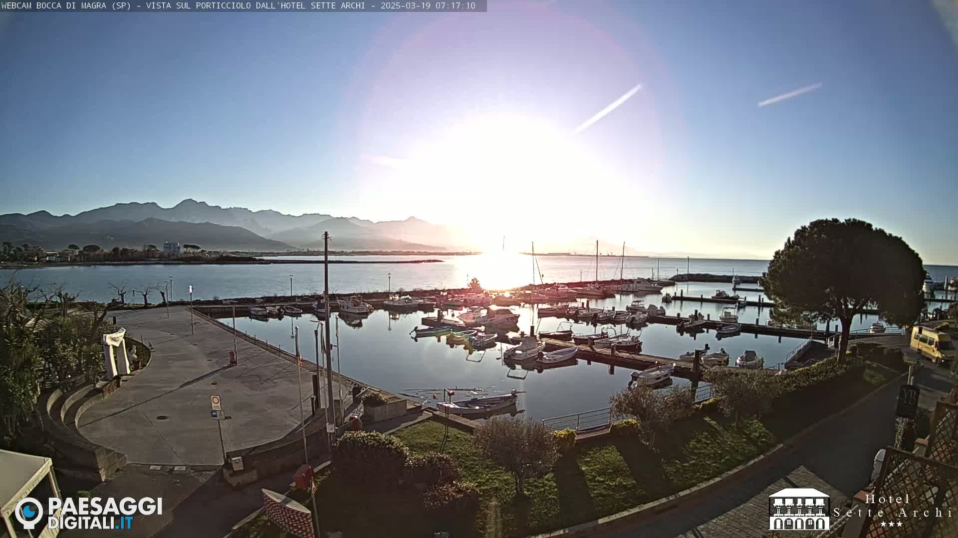 A marina filled with boats is seen at sunrise under a clear sky, with mountains in the distance.