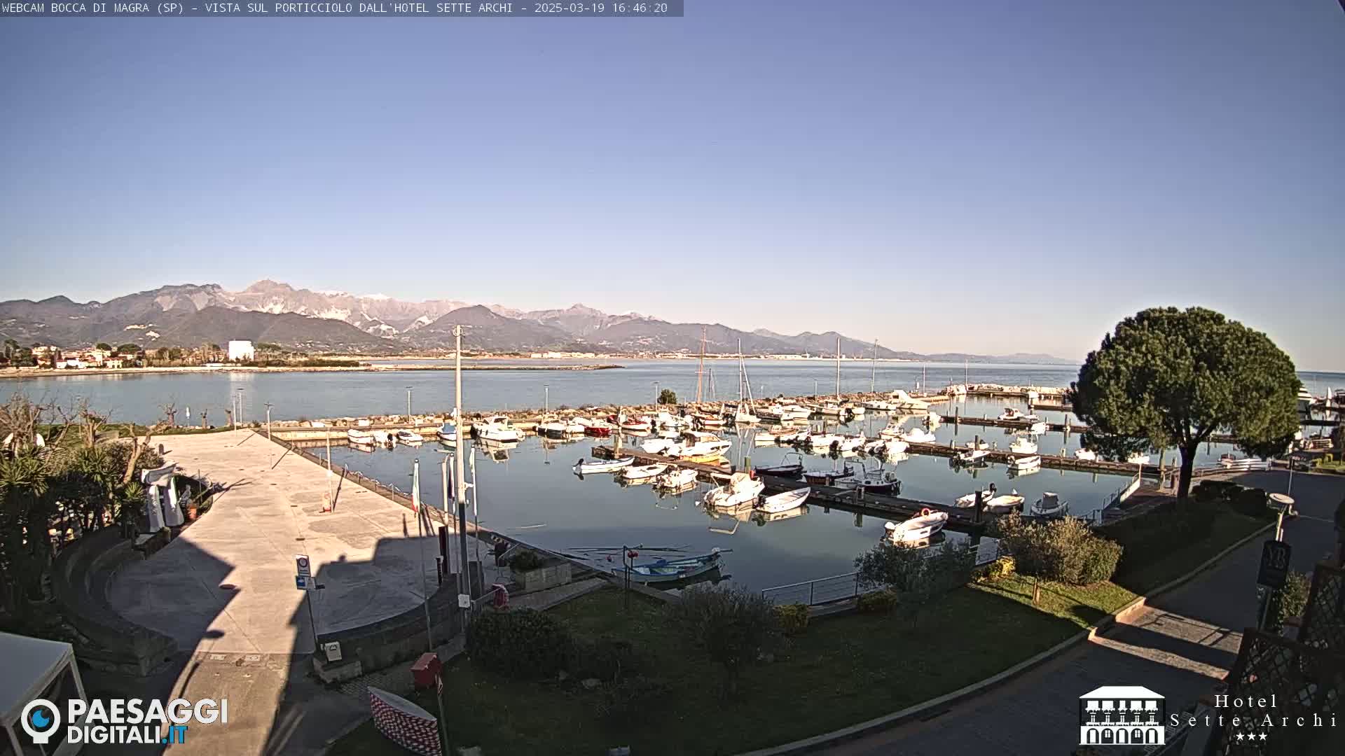 A marina filled with numerous boats is situated in front of a calm body of water, with mountains in the background under a clear sky.