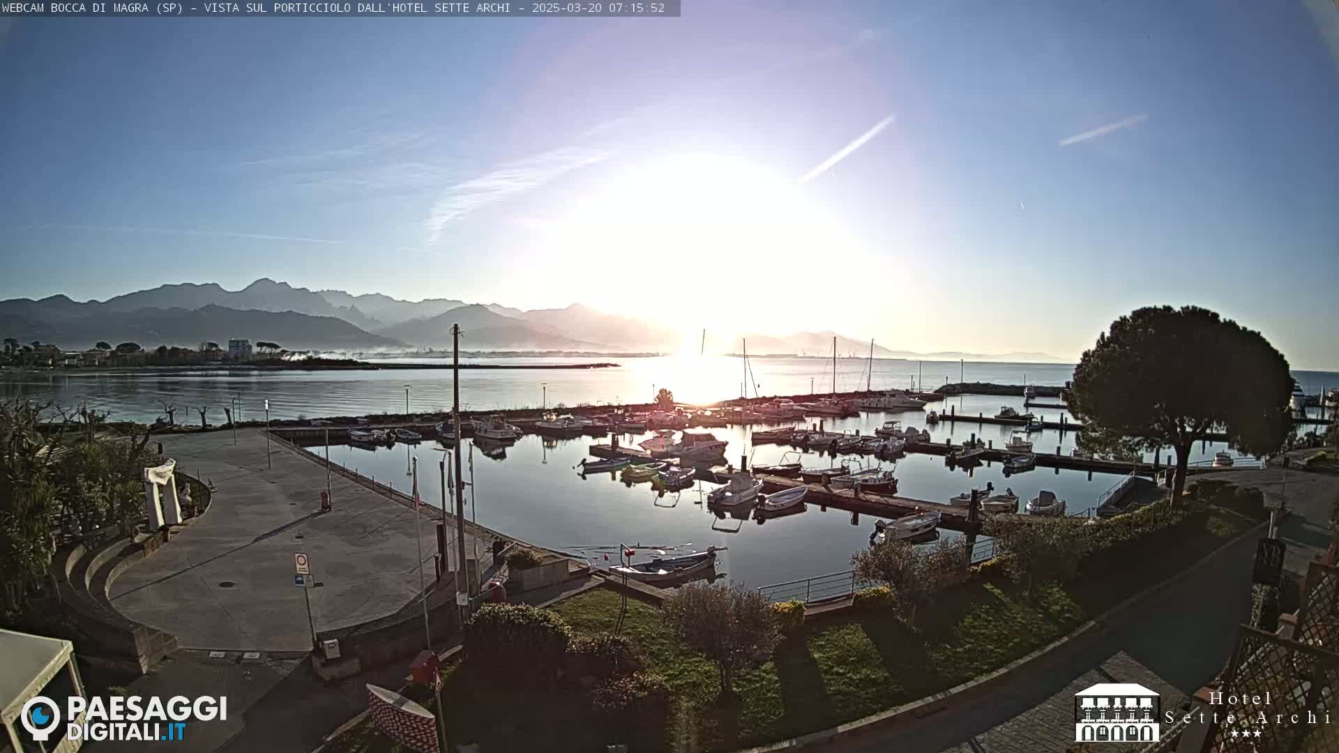 A marina filled with boats is seen at sunrise, with mountains in the distance under a clear sky.