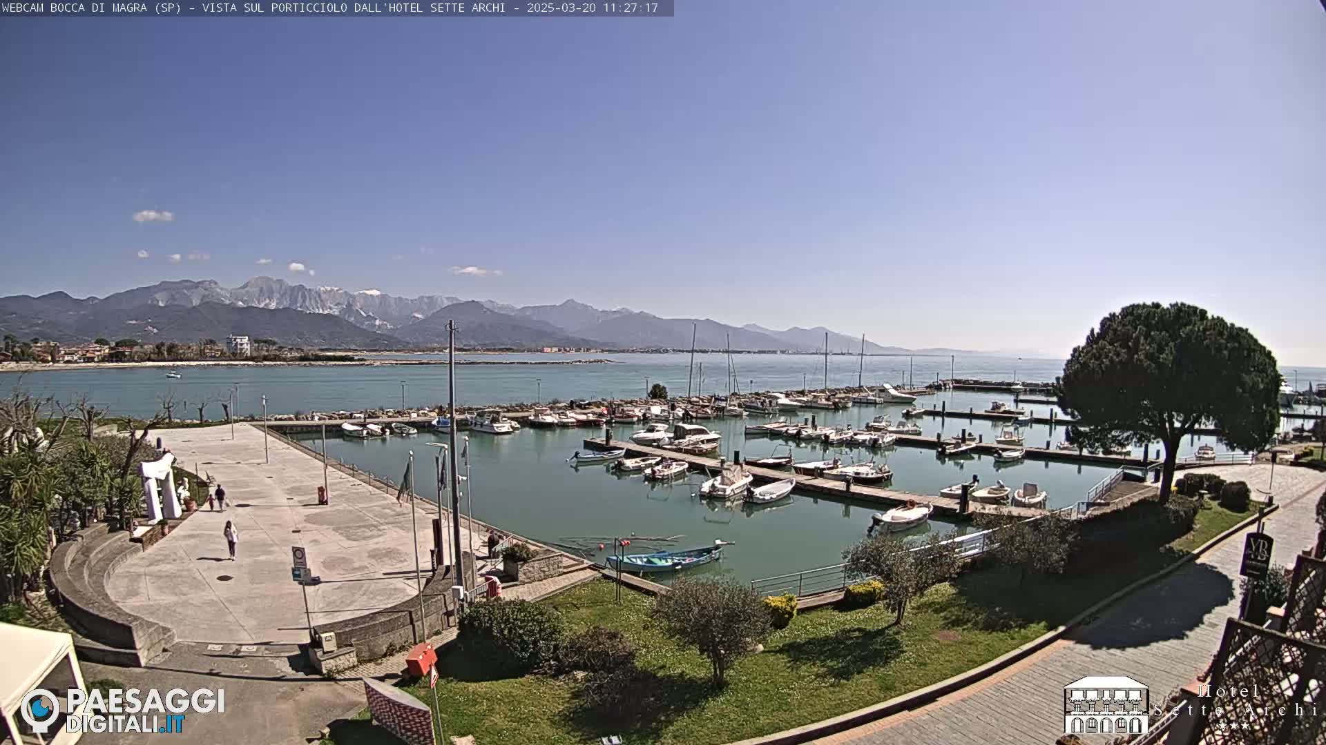 A marina filled with boats is situated next to a paved walkway, with a clear blue sky and mountains in the distance.