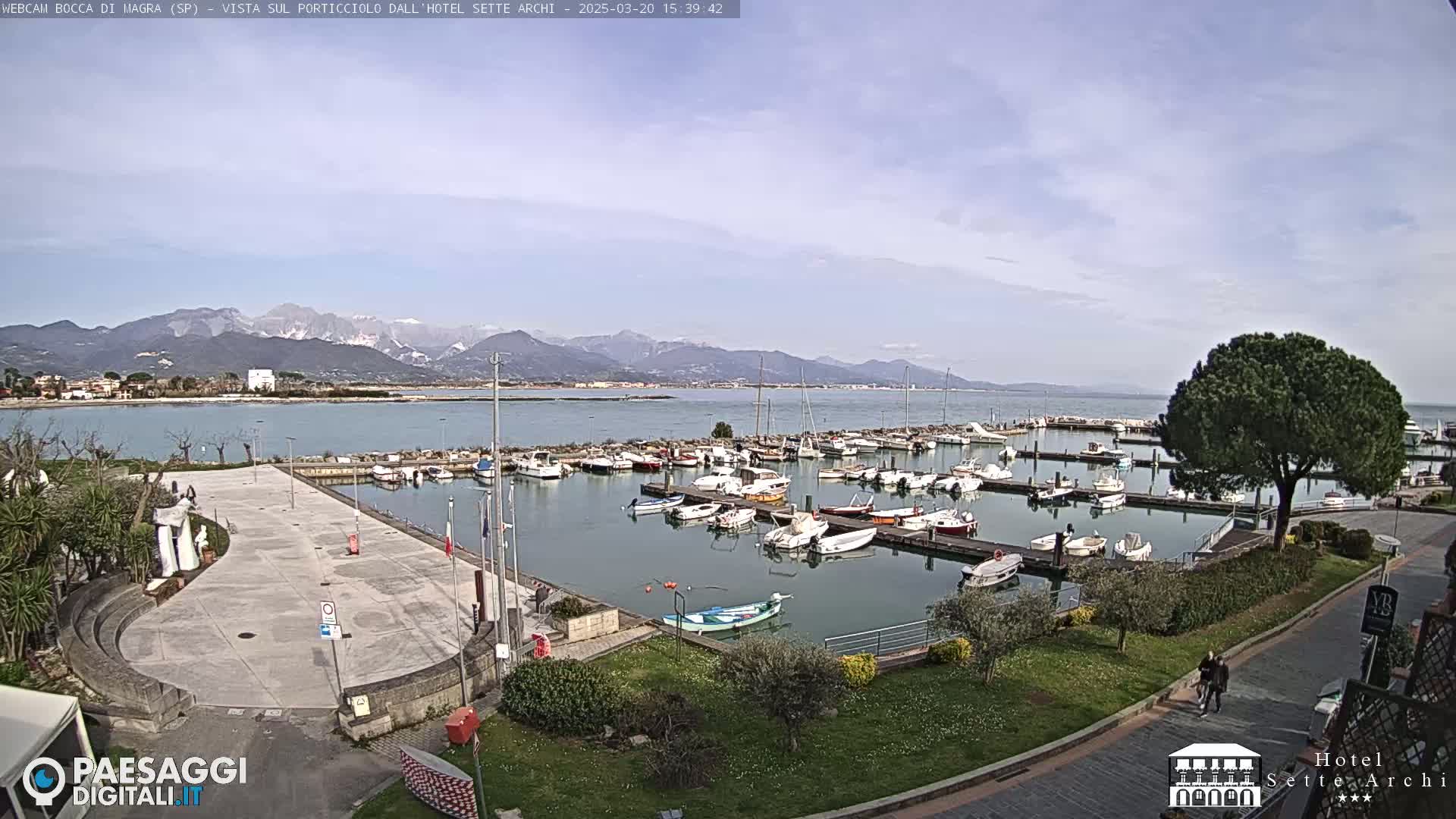 A marina filled with boats is situated near a paved plaza and green space, with snow-capped mountains visible in the background under a mostly clear sky.