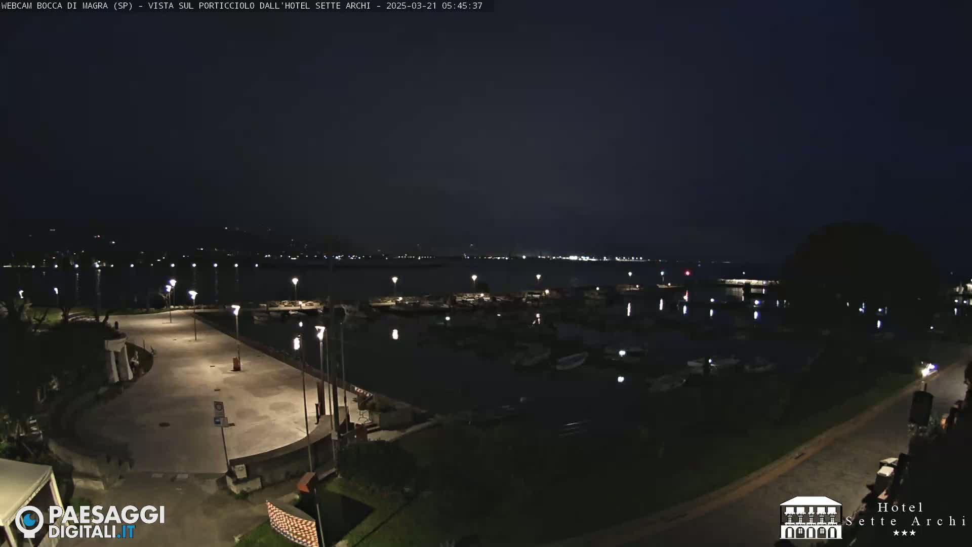 A nighttime view of a harbor filled with boats, a paved waterfront area with lights, and a dark, calm body of water extending to a dimly lit shoreline in the distance.