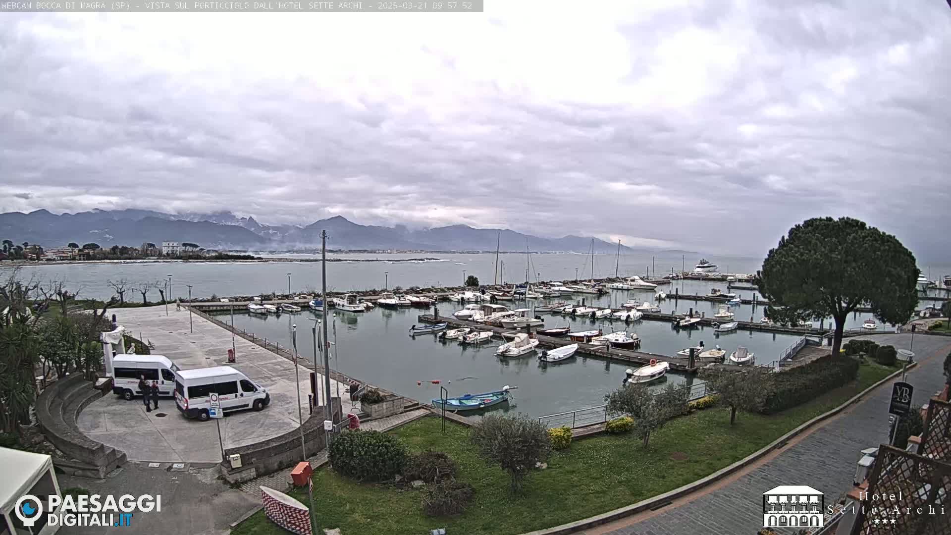 A marina filled with numerous boats is situated near a calm body of water, under a cloudy sky, with mountains visible in the distance.