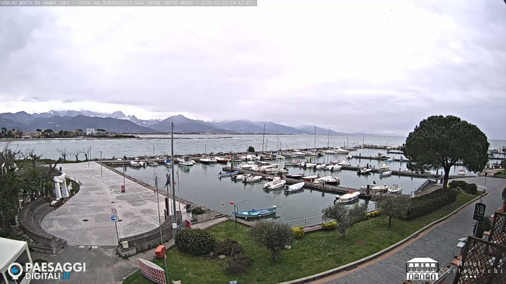 A harbor filled with boats is shown on an overcast day with mountains in the background.